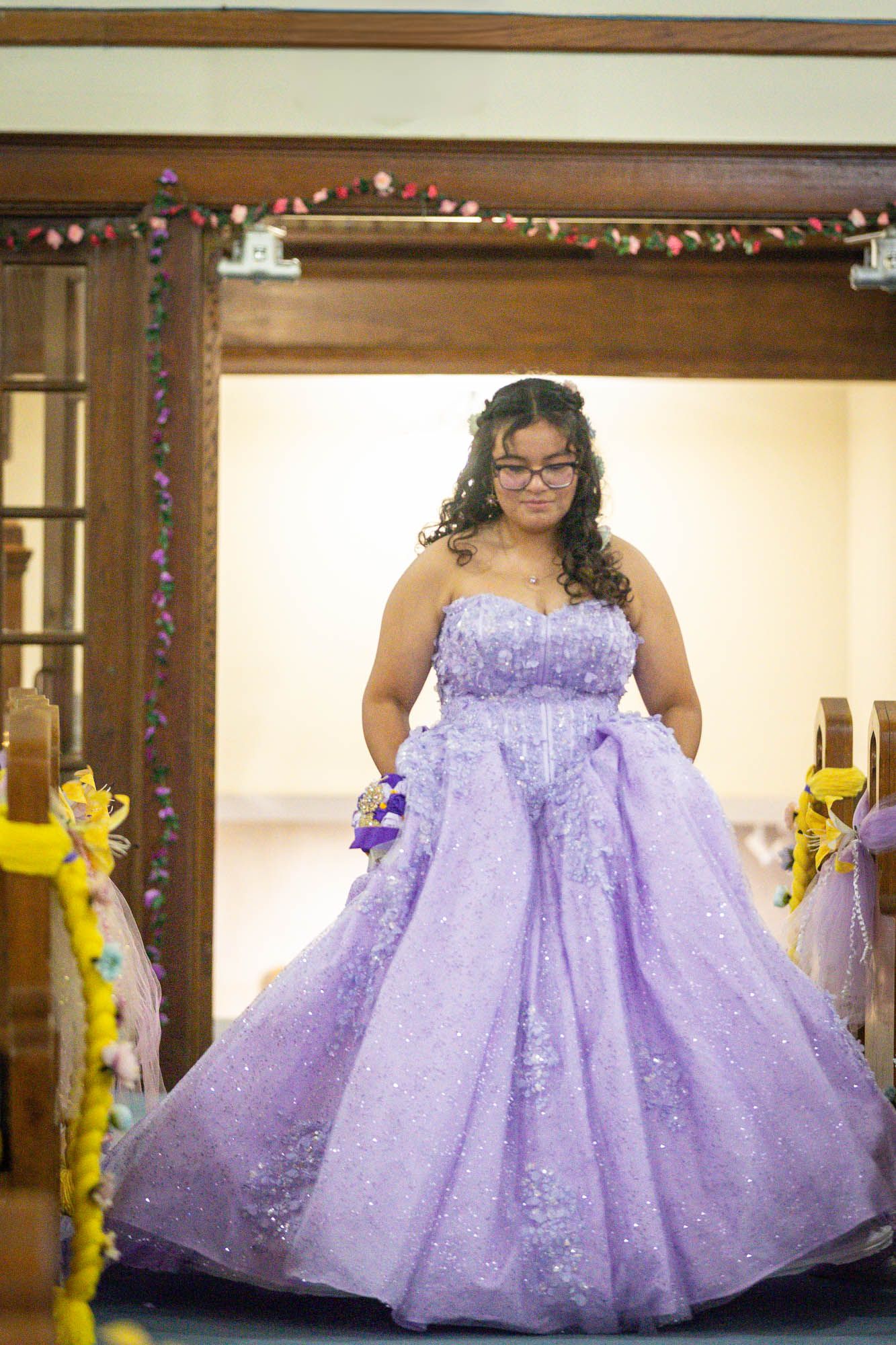 Young woman in a sparkling lavender strapless ball gown walks down a church aisle at a quinceañera.