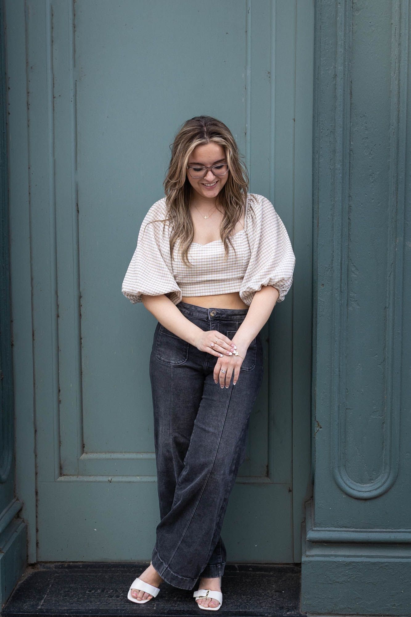 Stylish young woman in crop top and dark wide-leg jeans leaning against teal painted door.