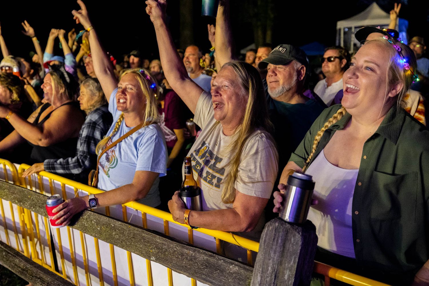 Excited crowd of fans cheering at outdoor concert barrier, woman raising arms in joy.