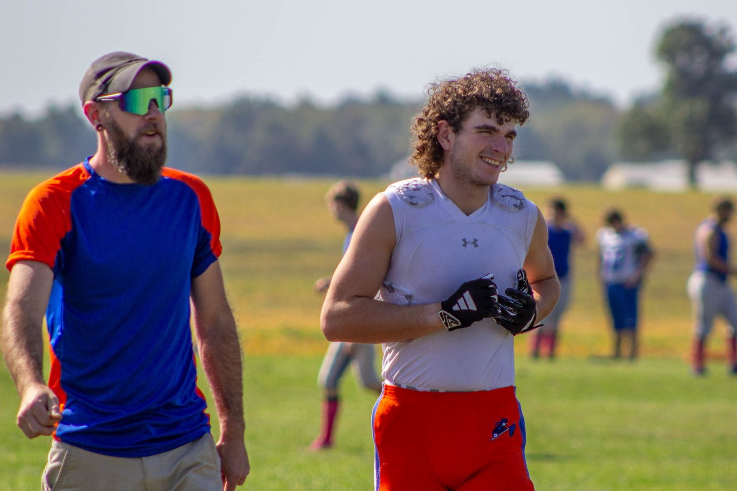 Football coach in blue shirt walks alongside curly-haired player in white jersey during outdoor practice.