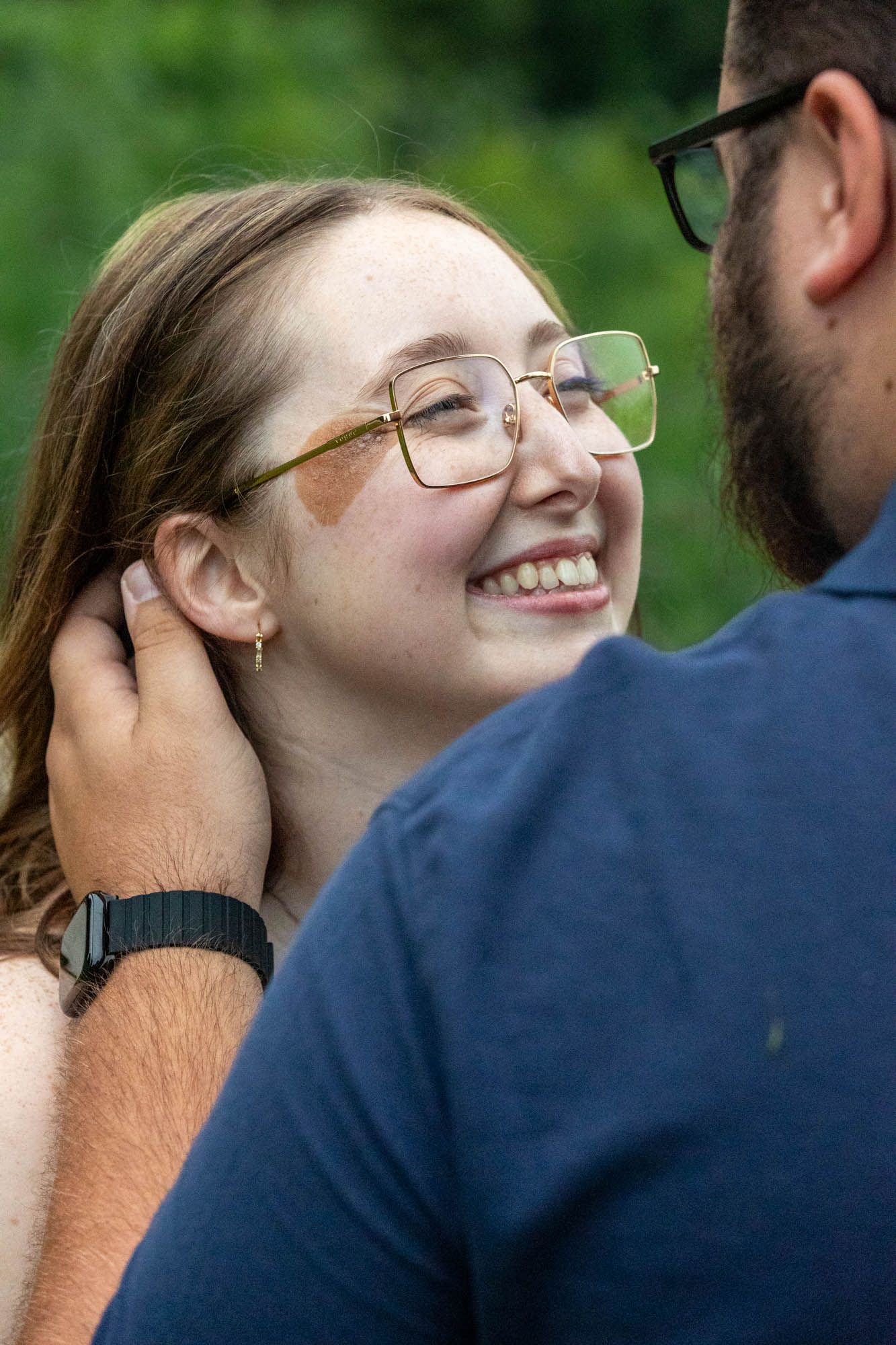Young woman with glasses laughing joyfully as man gently brushes hair from her face outdoors.