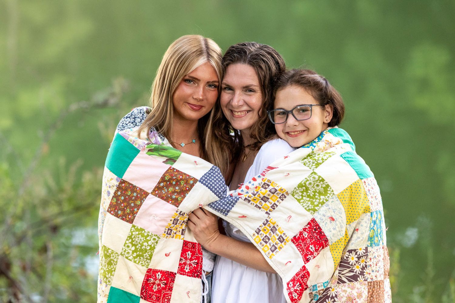 Three women wrapped in a colorful patchwork quilt smile together outdoors near a green, lush background.