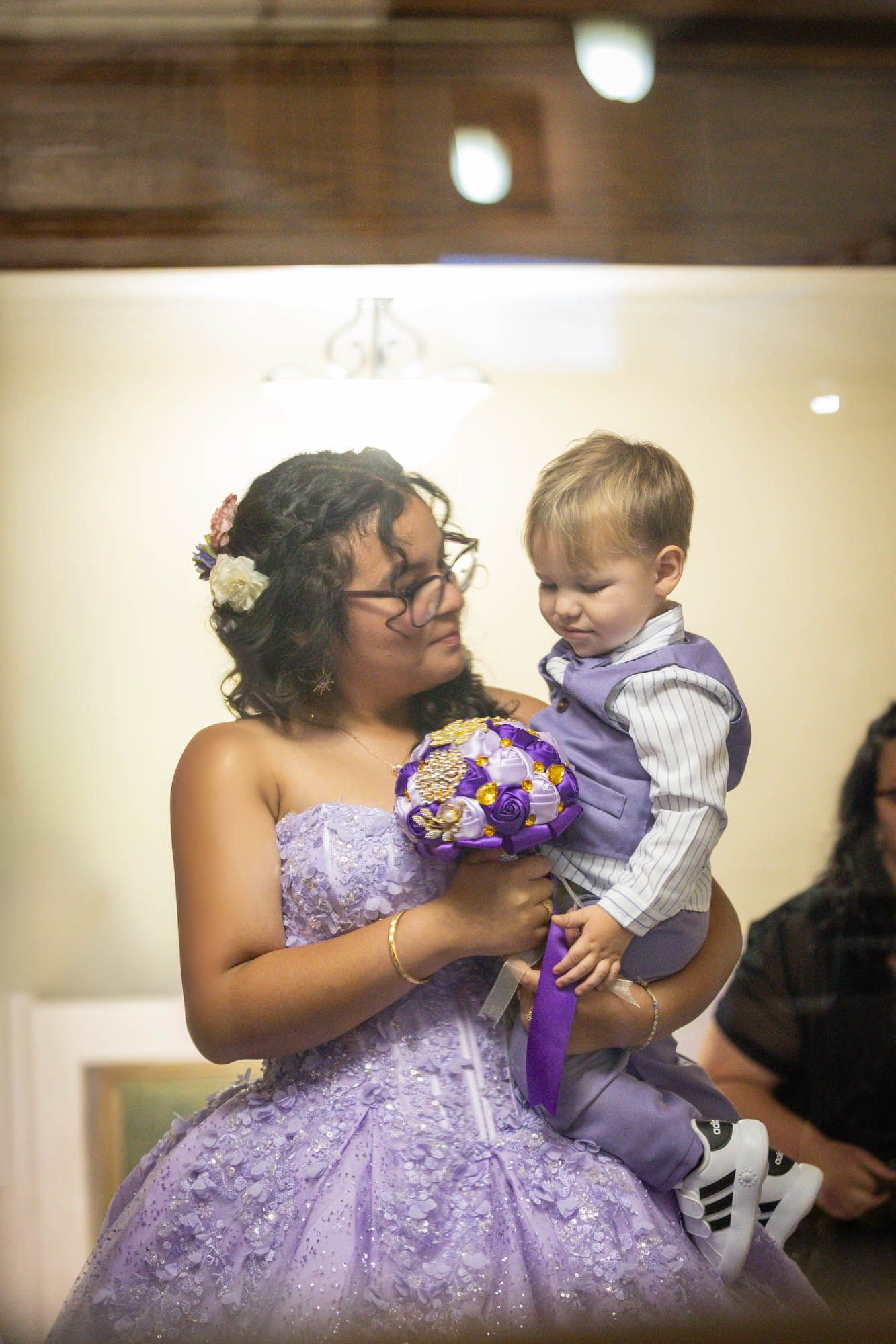 Quinceañera girl in lavender gown holding a baby boy in matching purple outfit during ceremony.