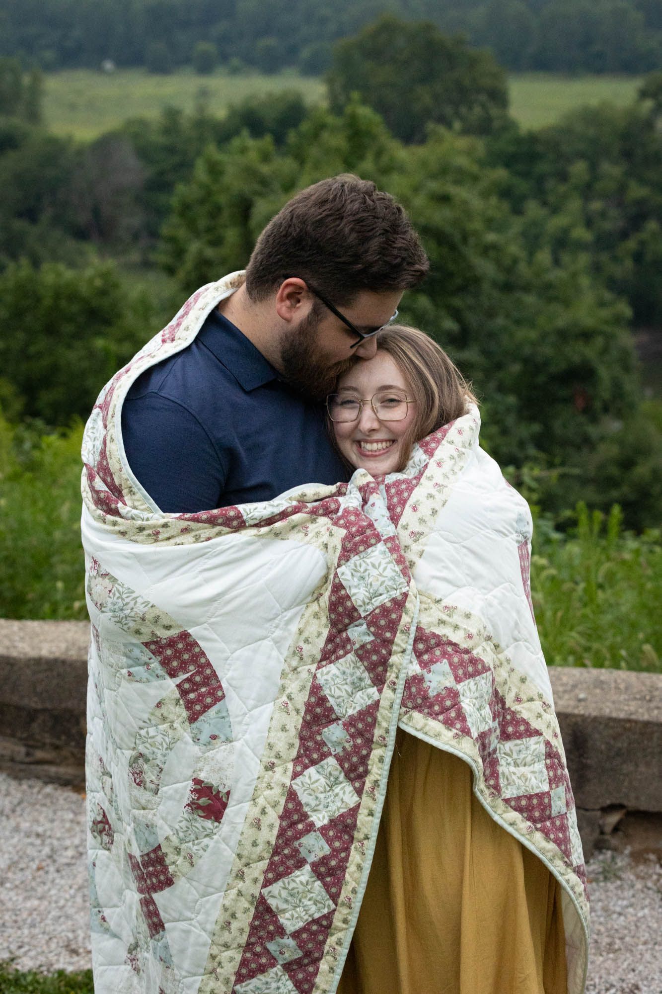 A couple wrapped in a patchwork quilt sharing a tender moment outdoors with lush green scenery behind them.