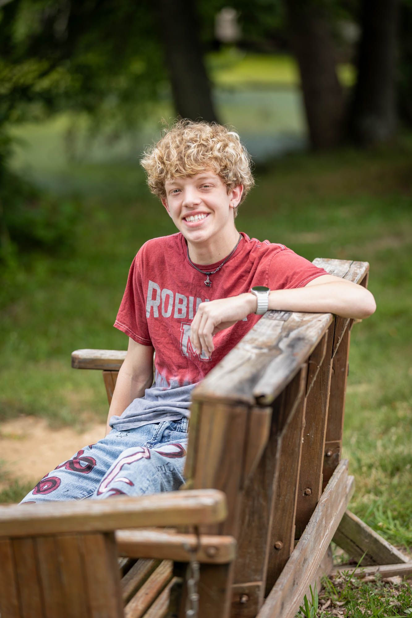 Smiling teenage boy with curly blond hair sitting on a wooden bench outdoors wearing a red Robin t-shirt.