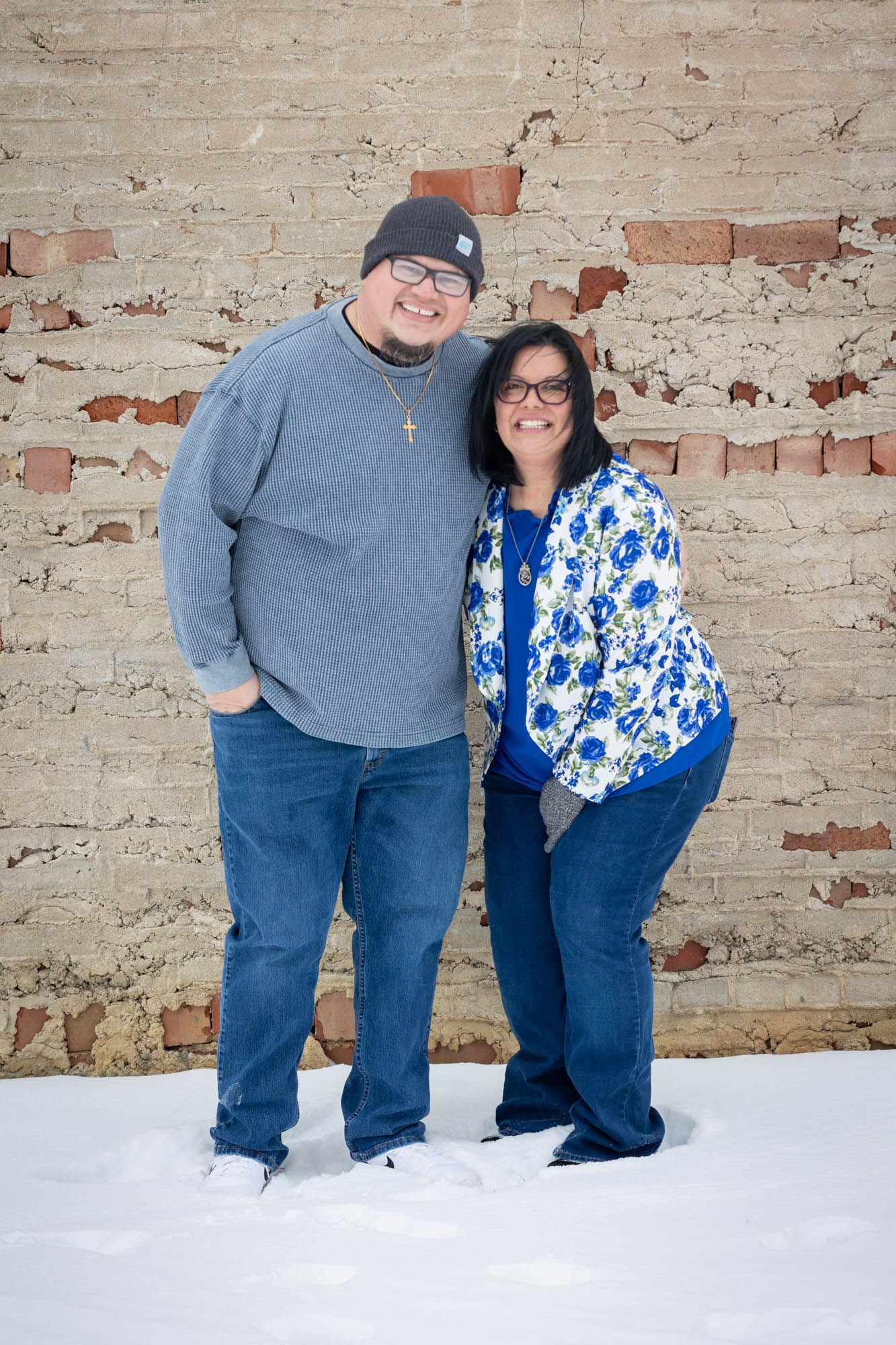 Couple smiling together in snow against rustic brick wall, woman in blue floral jacket and man in gray sweater with cross necklace.