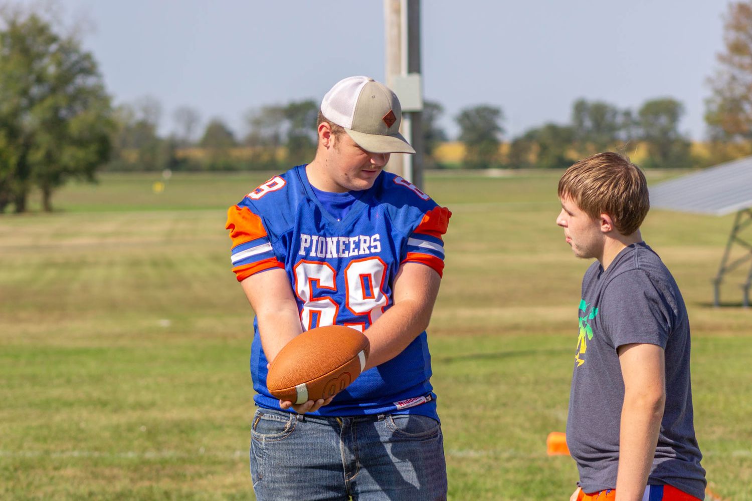 Pioneer football player in blue jersey shows a football to a younger player on a practice field.