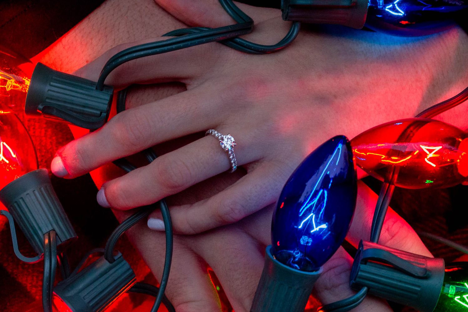 Couple holding hands adorned with diamond engagement ring surrounded by colorful Christmas lights.
