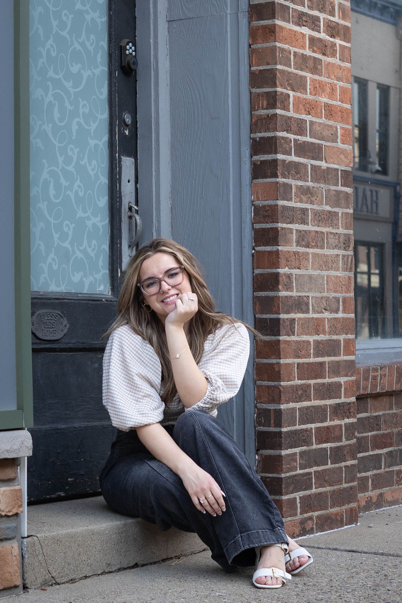 Happy young woman with glasses sitting on urban storefront steps, resting chin on hand and smiling.