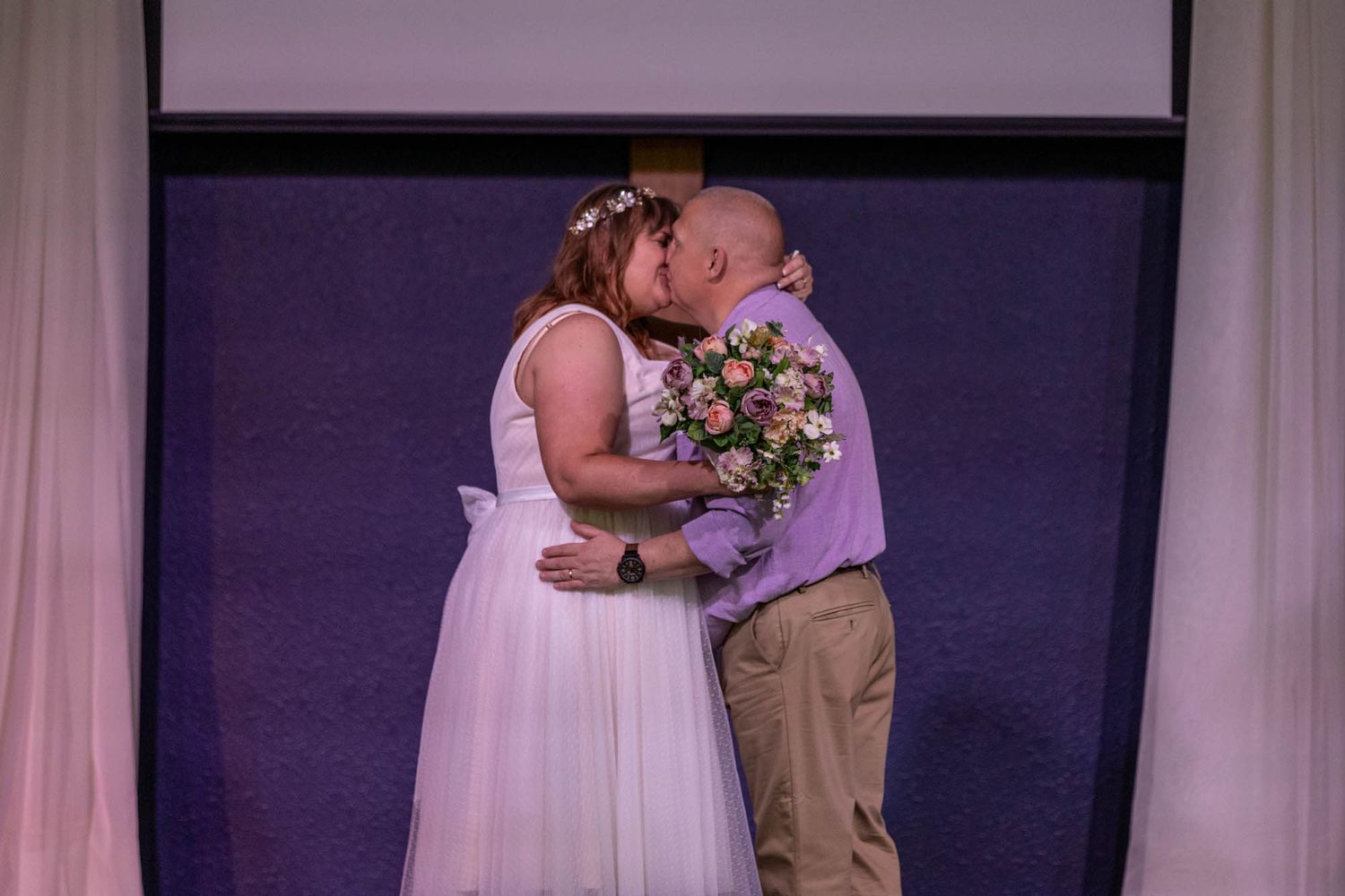 Bride and groom share a kiss at the altar, bride holding a pink bouquet in a white wedding dress.