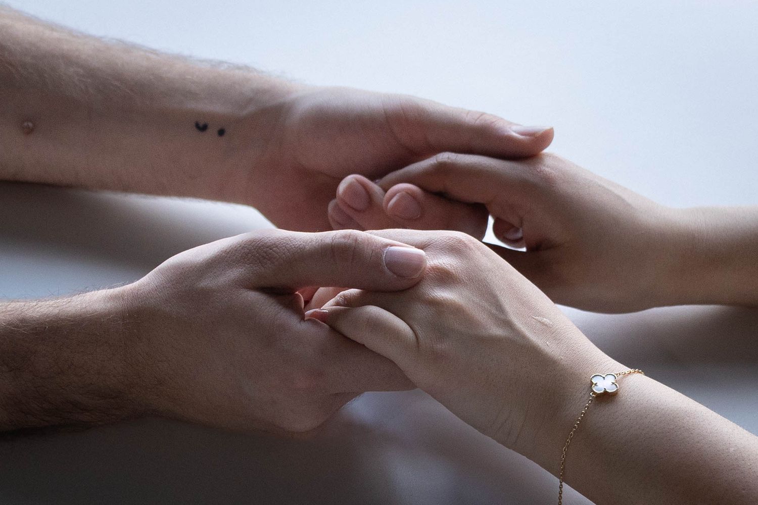 Two people holding hands tenderly, one with a small tattoo and the other wearing a delicate bracelet.