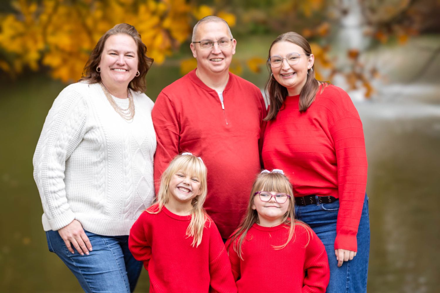 Family of five smiling together outdoors in autumn, wearing red and white near a waterfall backdrop.