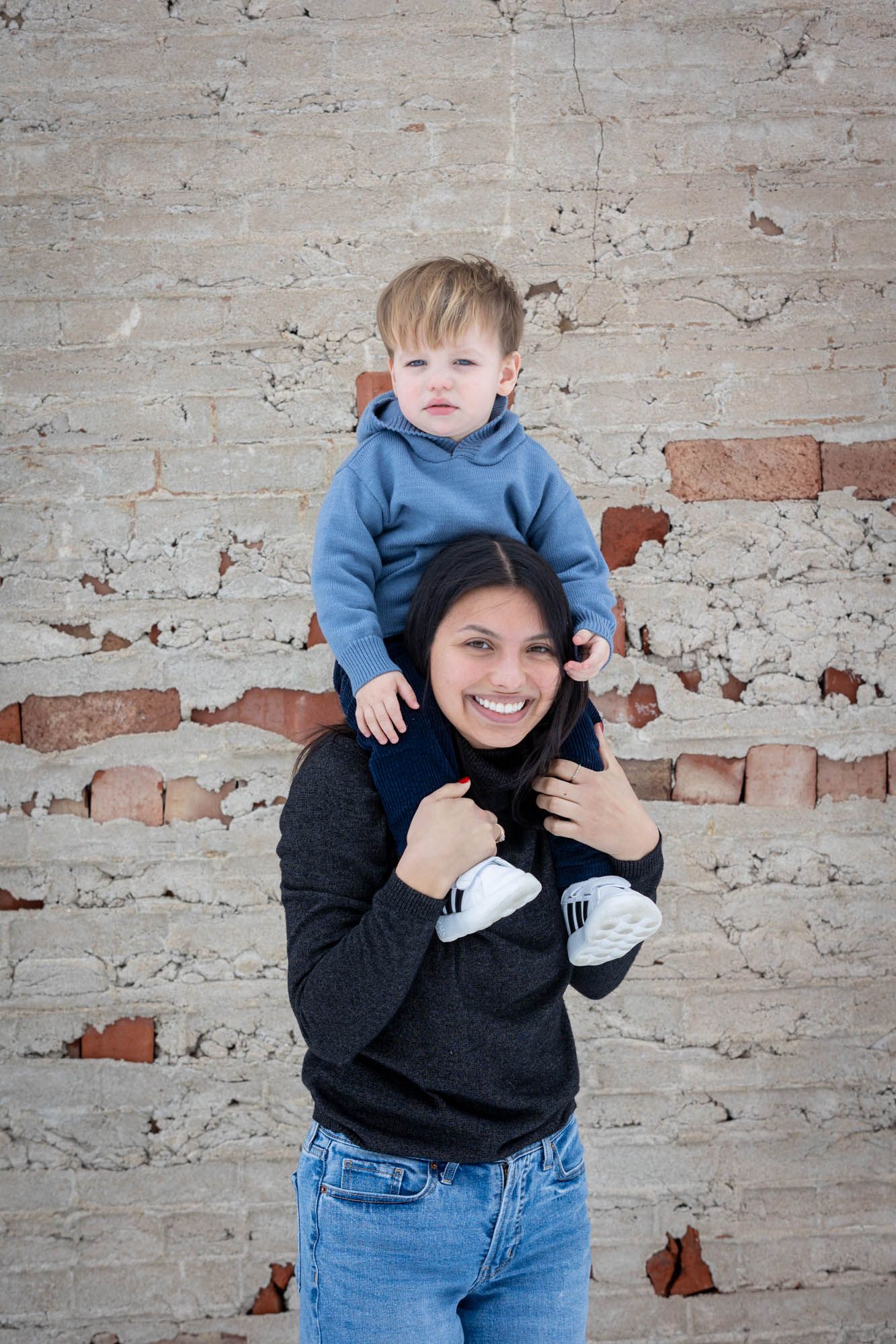 Smiling young woman carrying a toddler boy on her shoulders in front of a rustic brick wall.