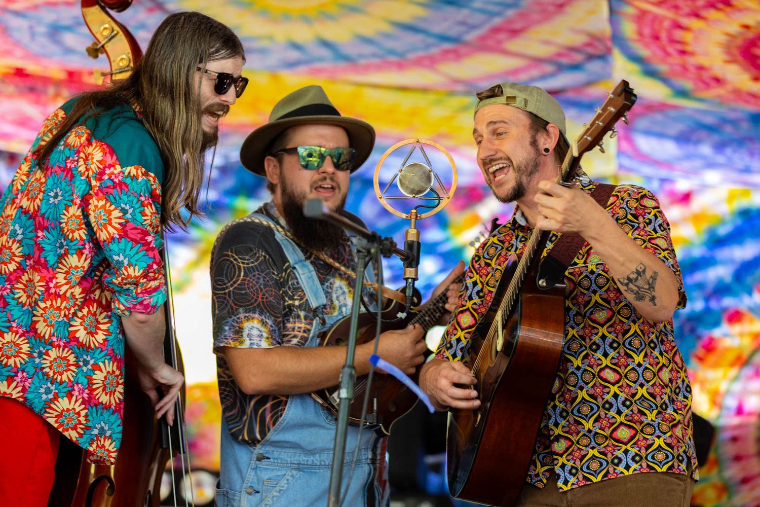 Three male musicians laugh and perform together on stage at a vibrant outdoor music festival.