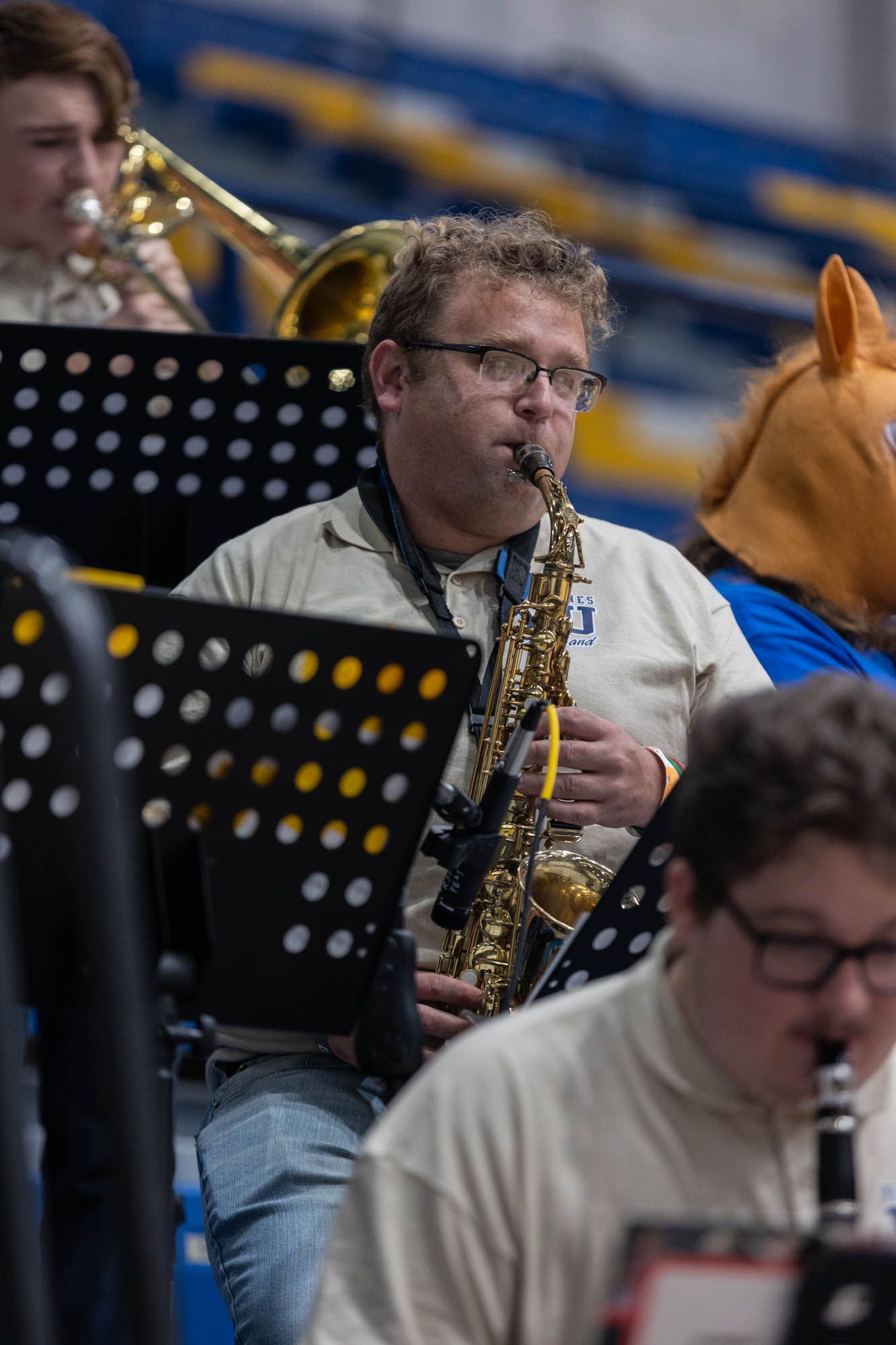 Saxophone player in glasses performs with college pep band during indoor basketball game in gymnasium.