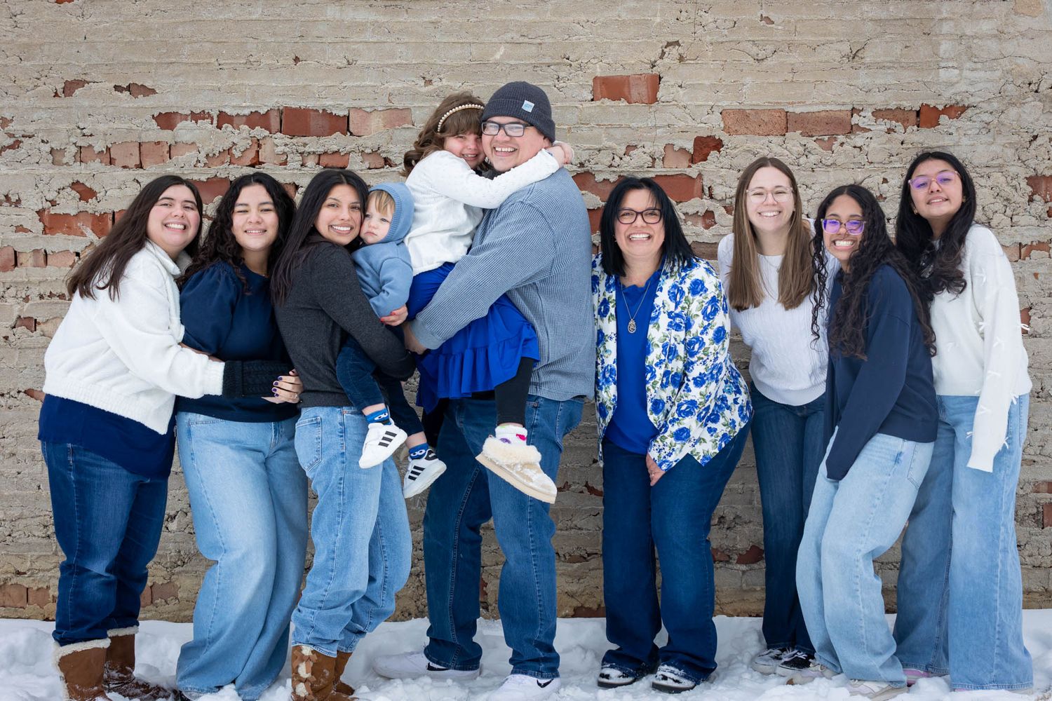 Large Hispanic family posing together in winter snow against rustic brick wall, smiling outdoors.
