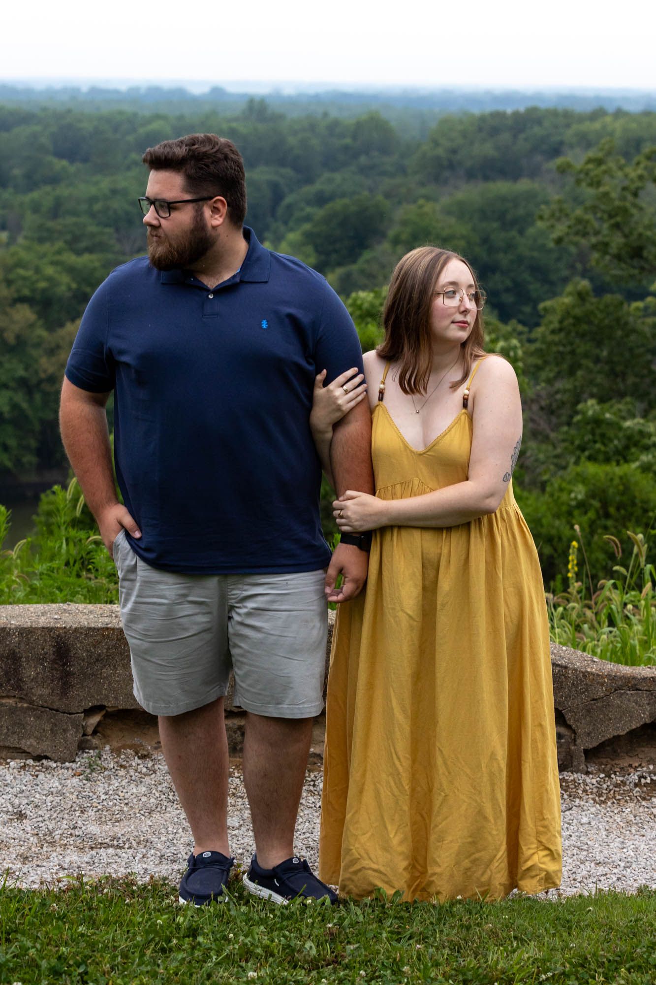 Couple standing outdoors at scenic overlook, man in blue polo and woman in yellow maxi dress, green trees background.