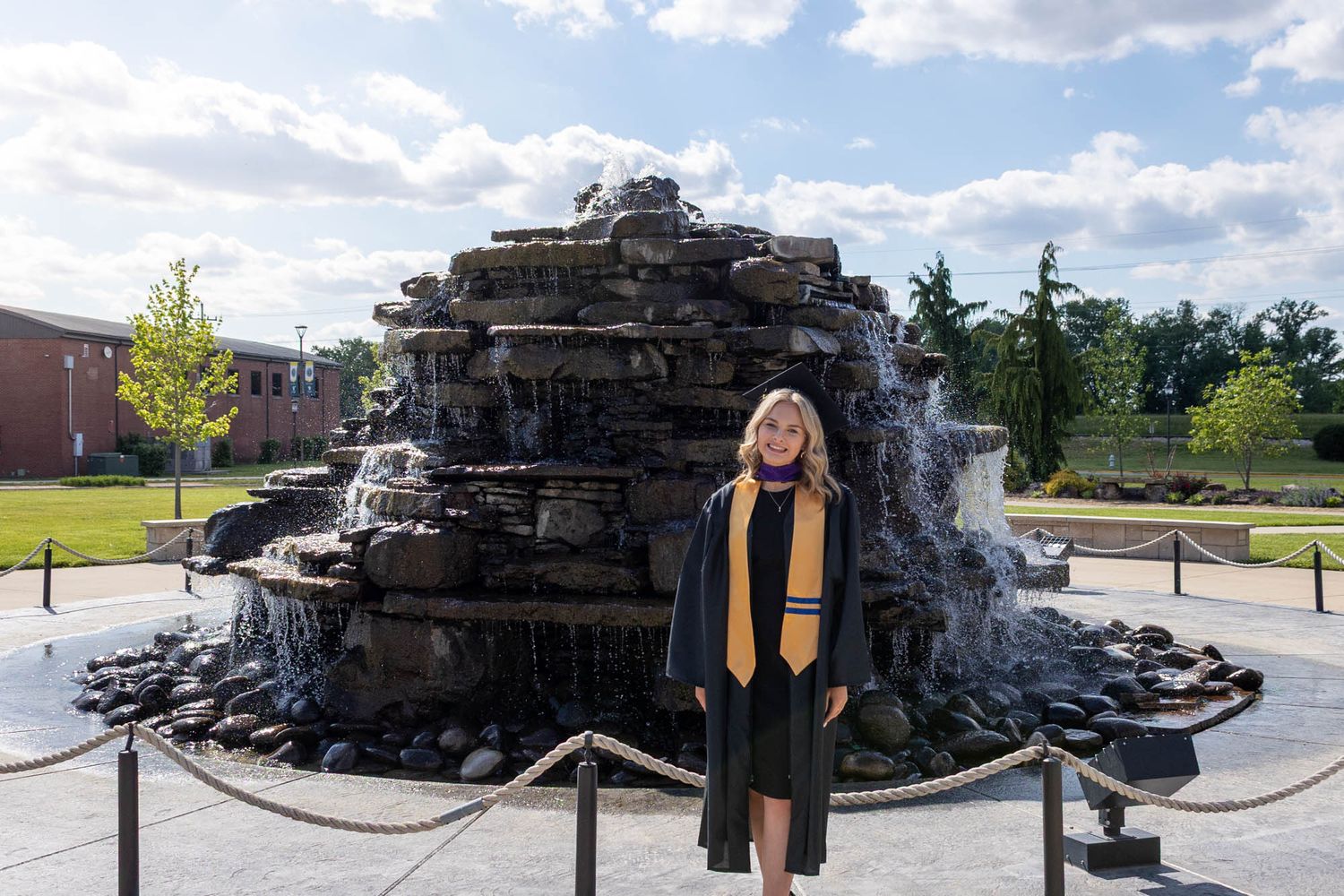 Graduate in cap and gown poses before a large rock waterfall fountain on a sunny college campus.