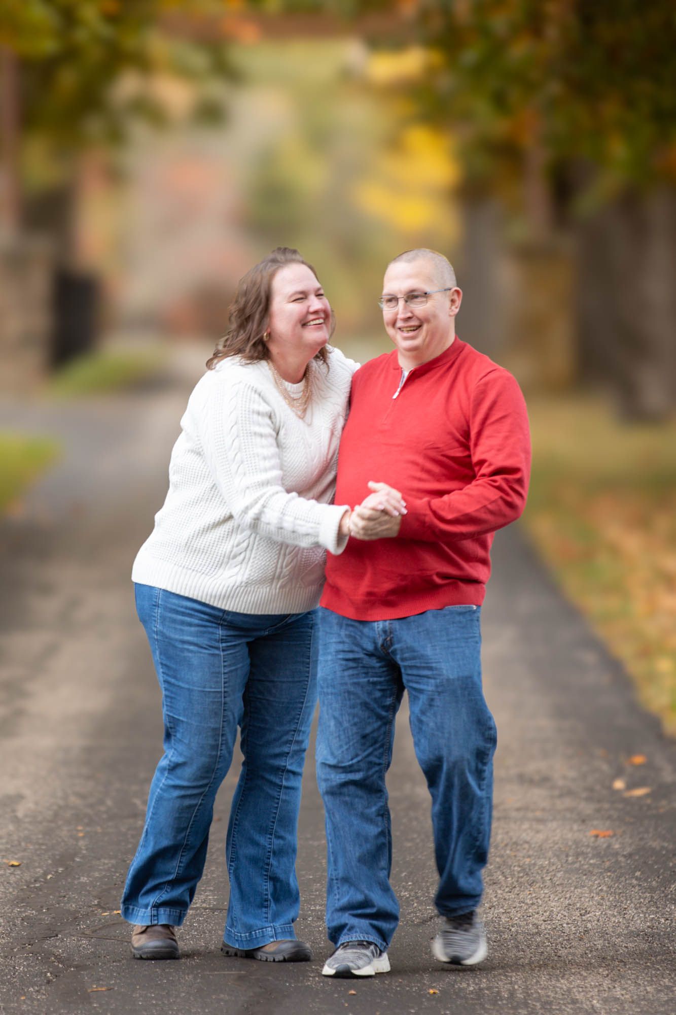 Laughing couple dances together on a scenic autumn road lined with colorful fall trees.