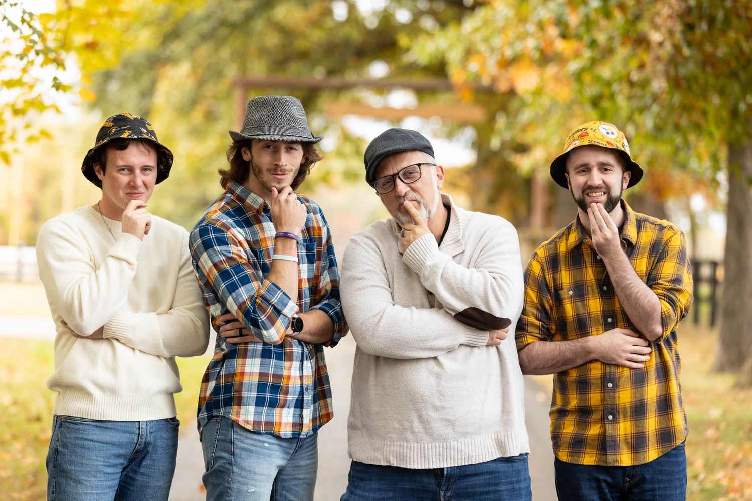 Four men wearing hats strike thoughtful poses together on a tree-lined autumn park path.