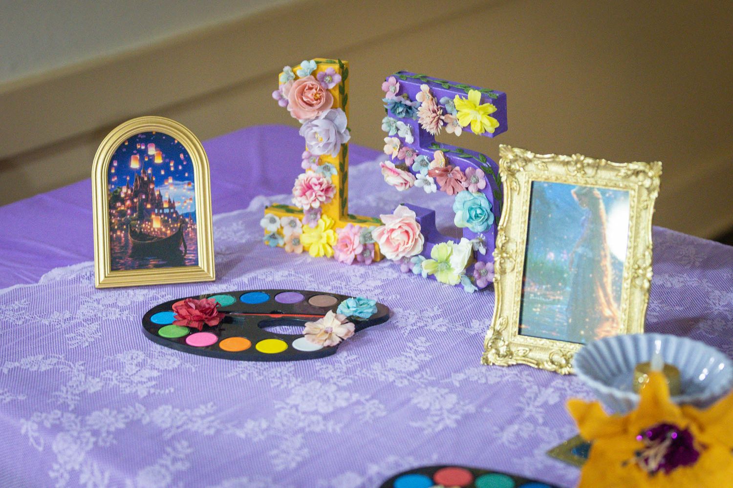 Rapunzel-themed party table with floral letters, lantern art, paint palette, and gold frame on purple lace cloth.