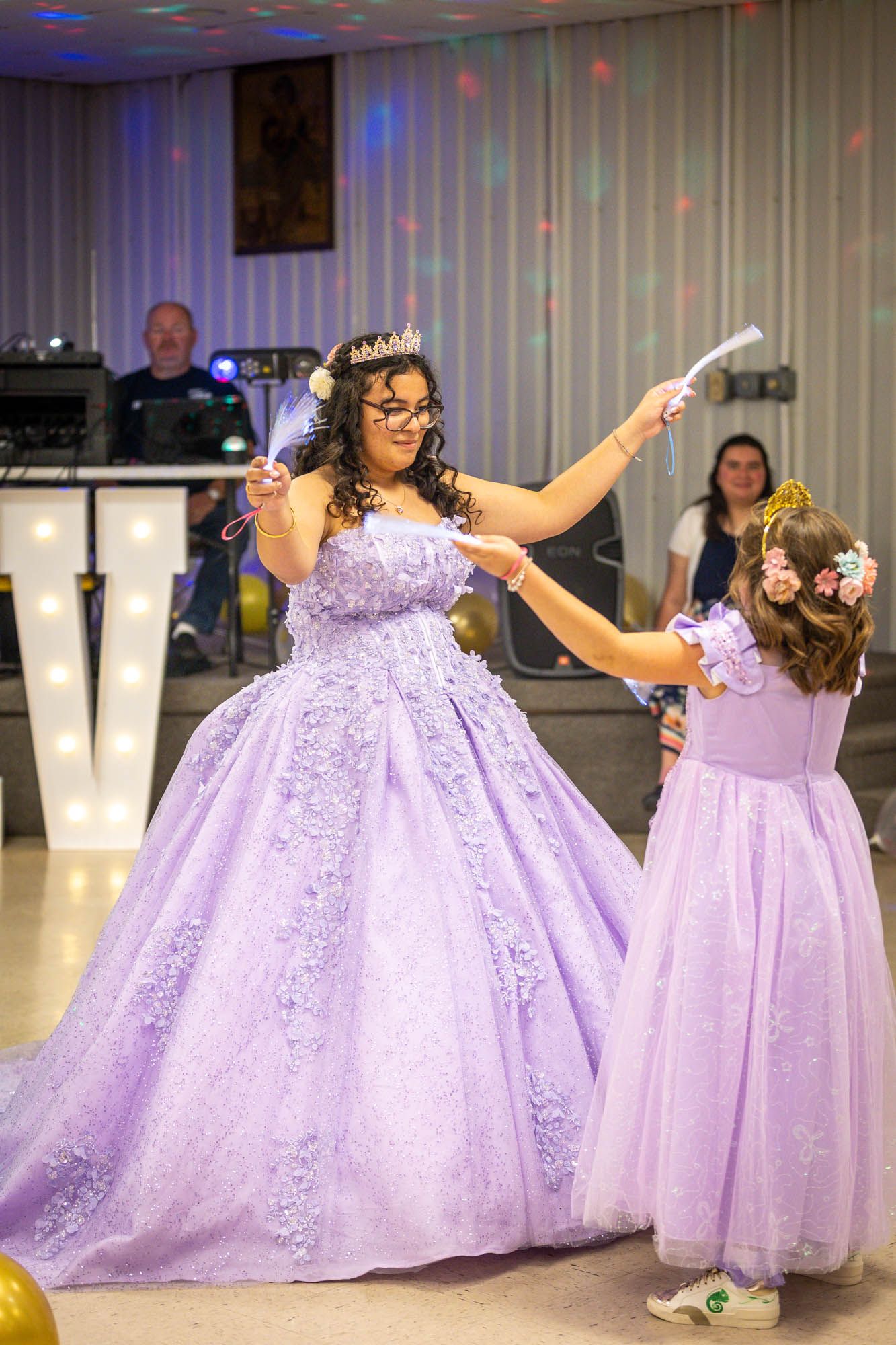 Teen in lavender ball gown dances with young girl in matching purple dress at quinceañera party.