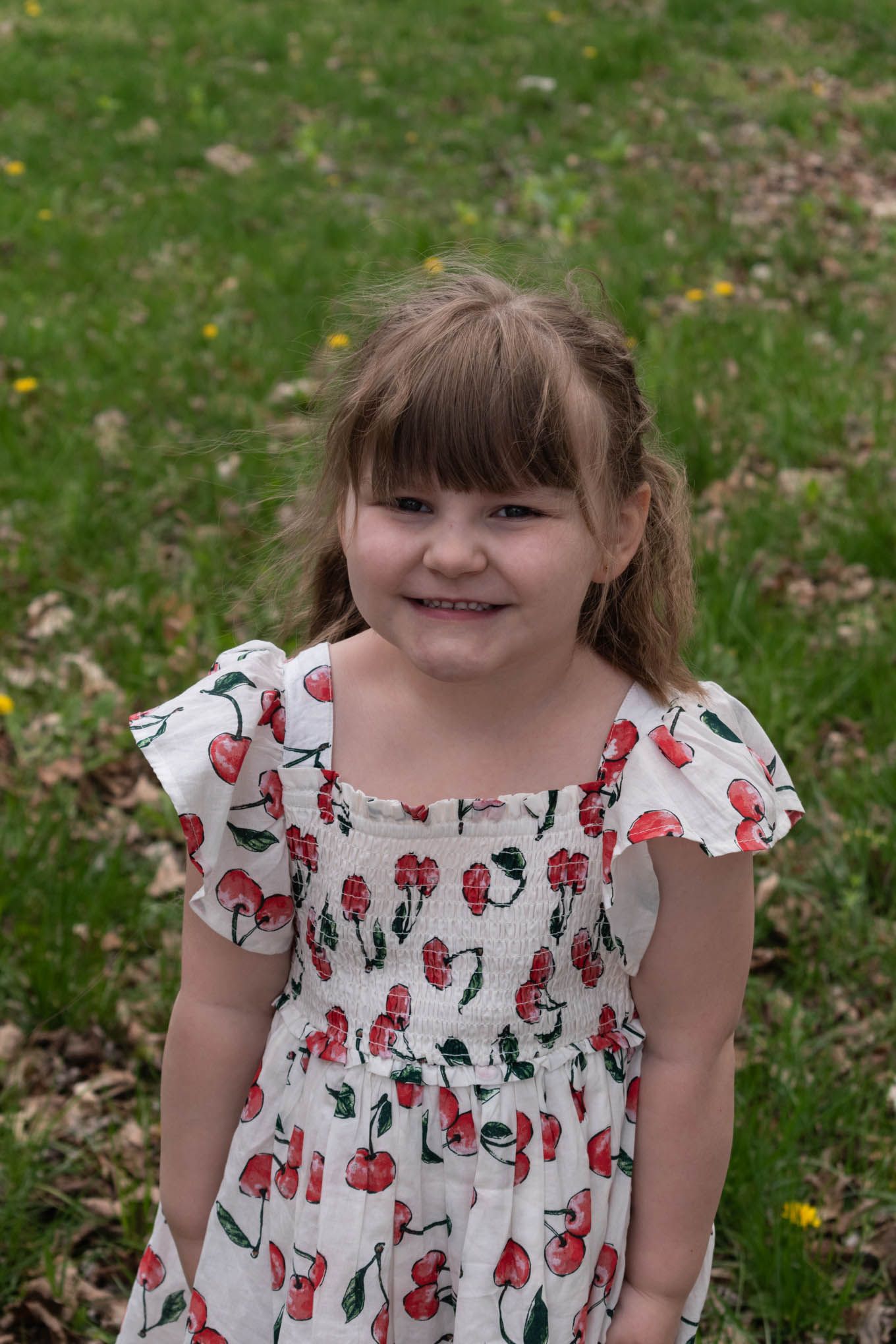 Adorable little girl in a cherry print dress smiles sweetly while posing outdoors on green grass.