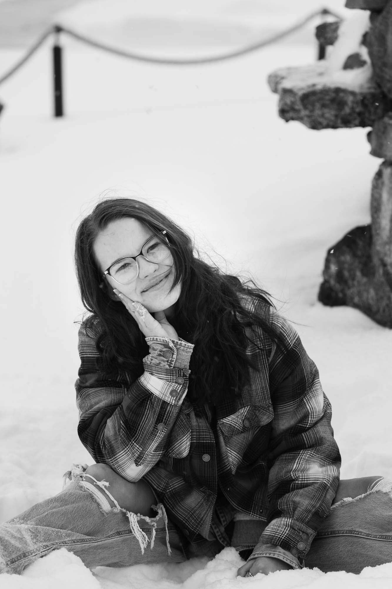Black and white portrait of a young woman with glasses sitting in snow, resting her chin on her hand.