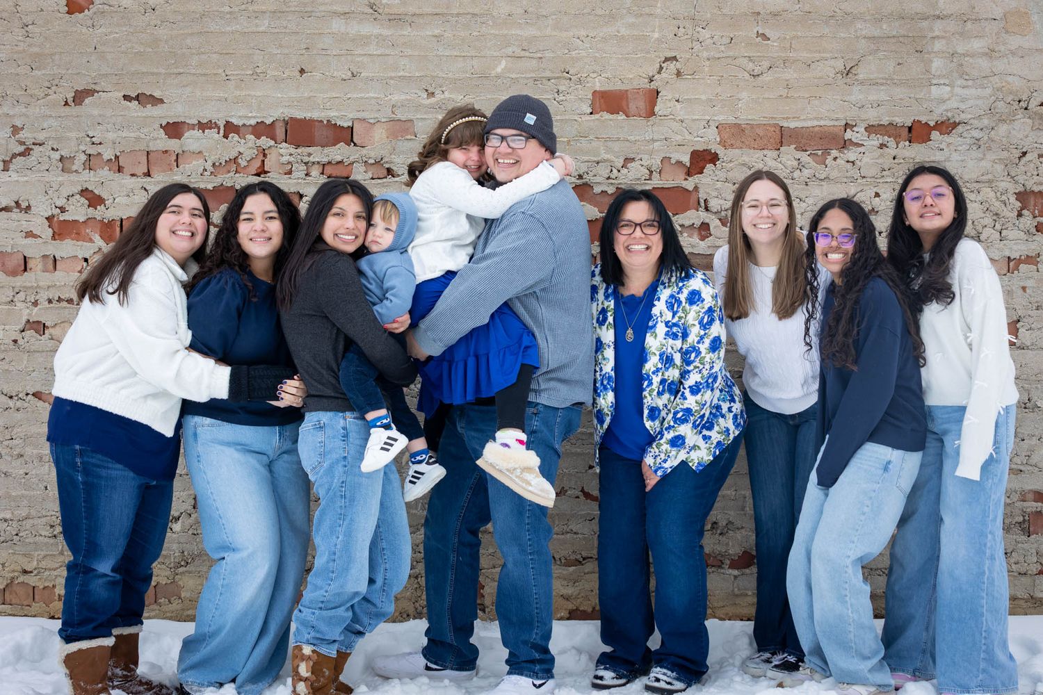 Large family group smiling together in winter, posing against a rustic brick wall with snow on the ground.