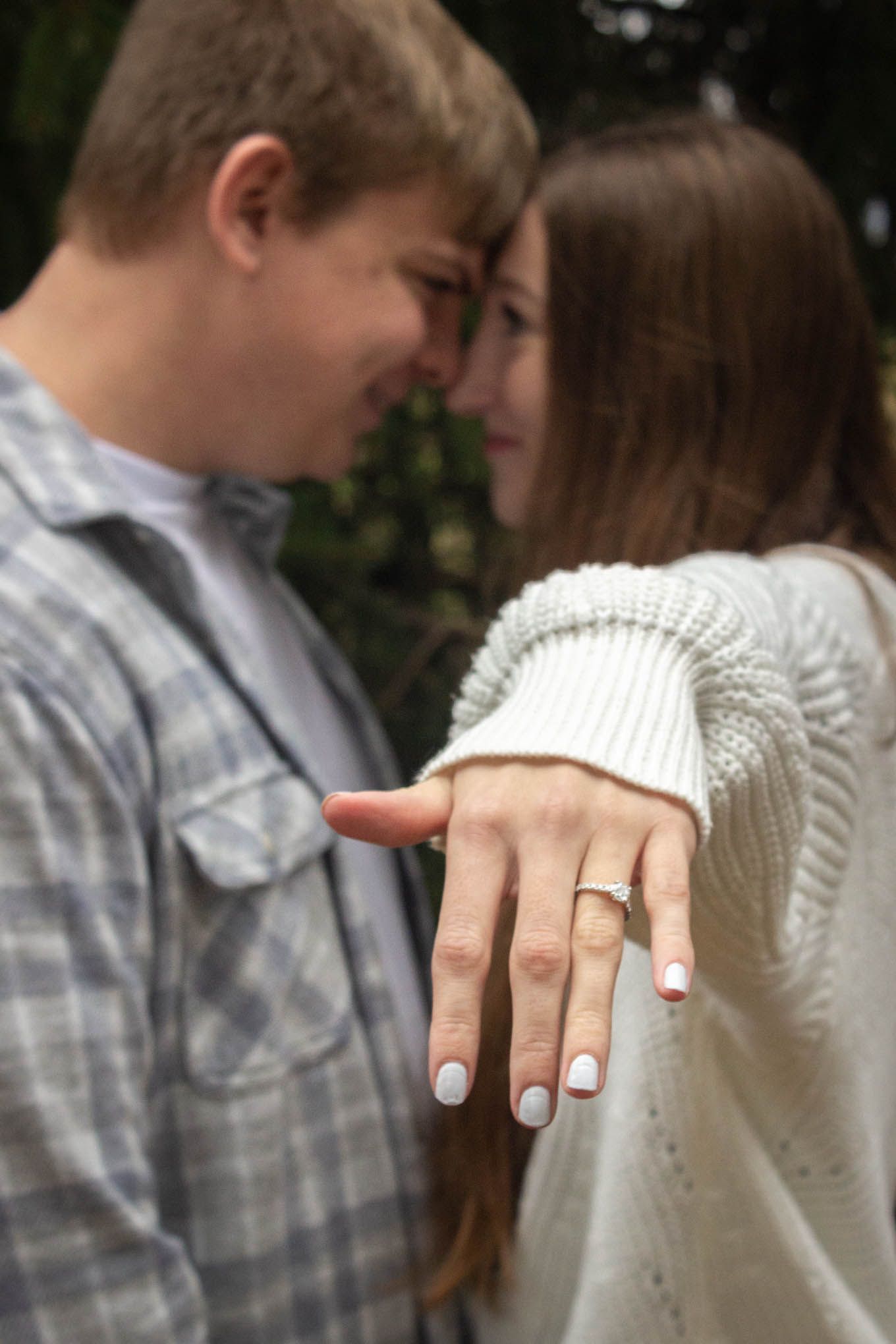 Couple facing each other as woman displays diamond engagement ring toward camera outdoors.