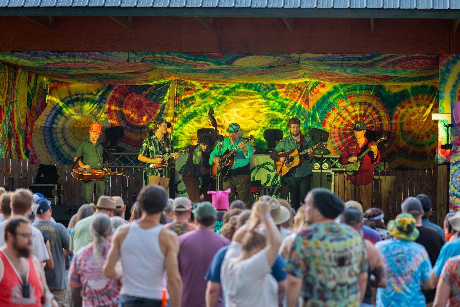 Live outdoor music festival with band performing on tie-dye decorated stage before large crowd.