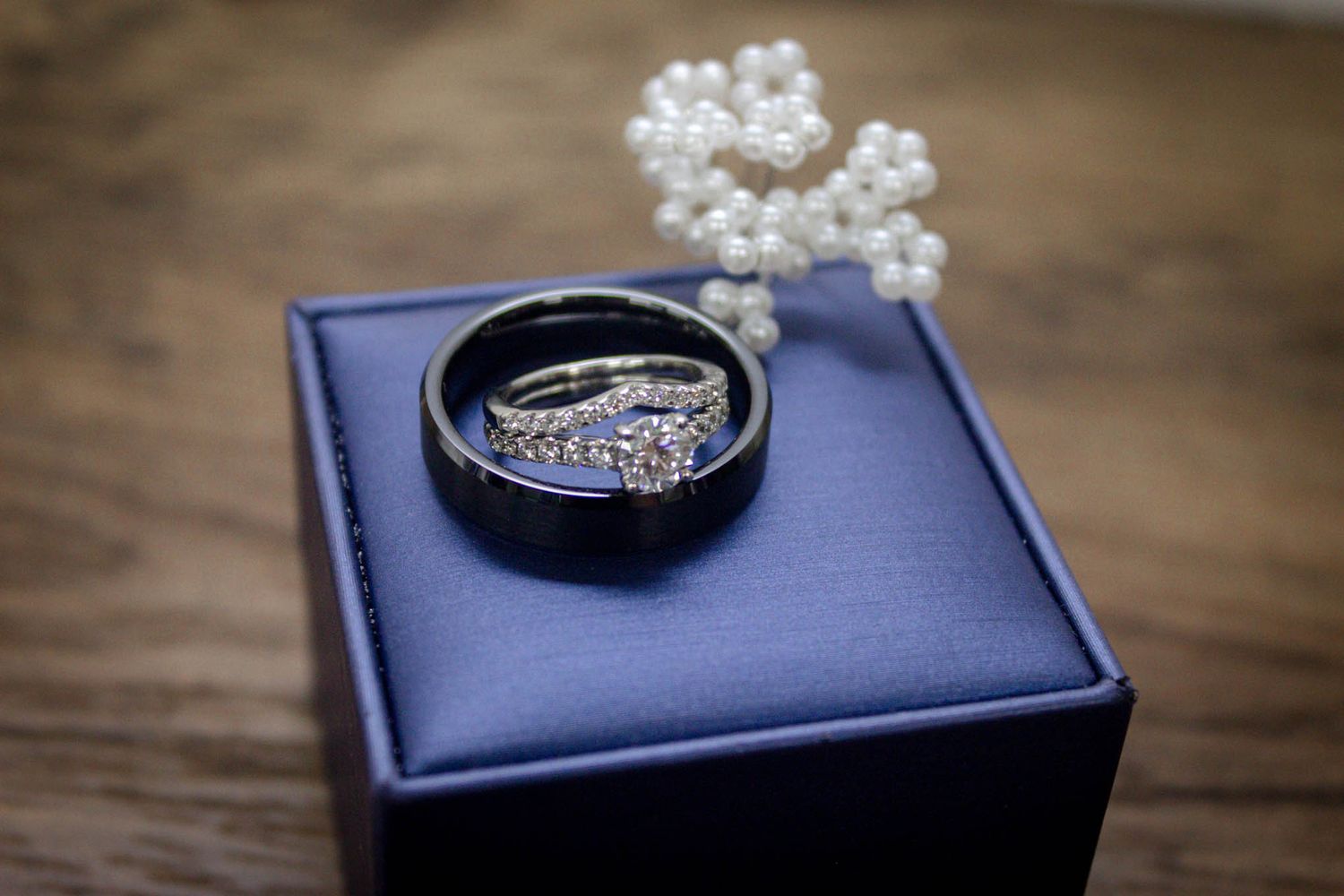 Wedding rings displayed on blue velvet box with pearl floral decoration on wooden surface.