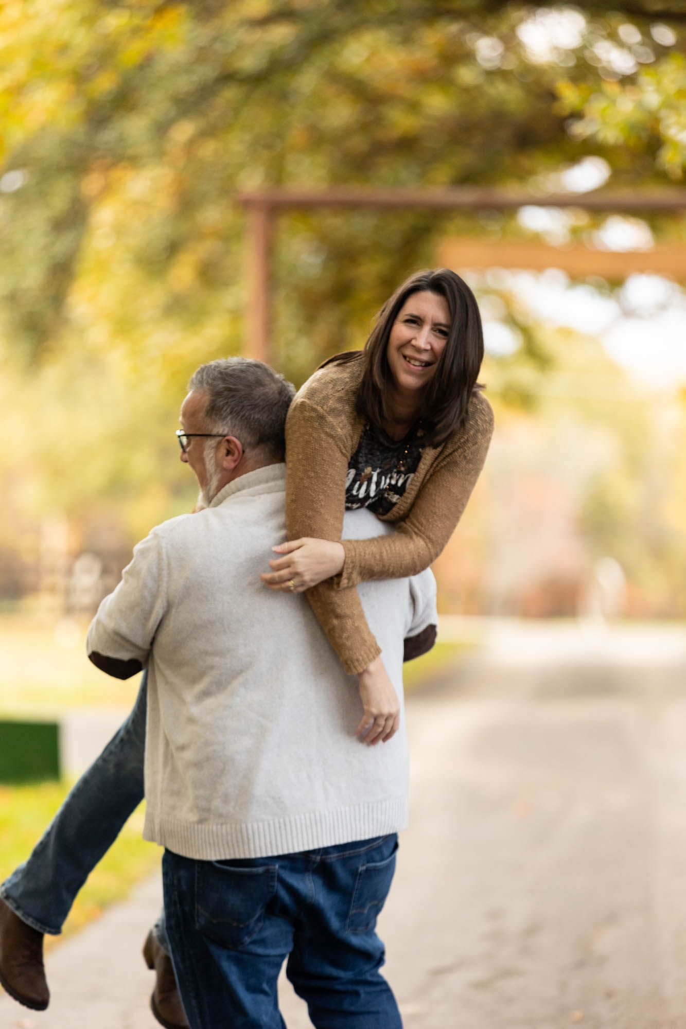 Man carries smiling woman in his arms along a tree-lined path during autumn engagement shoot.