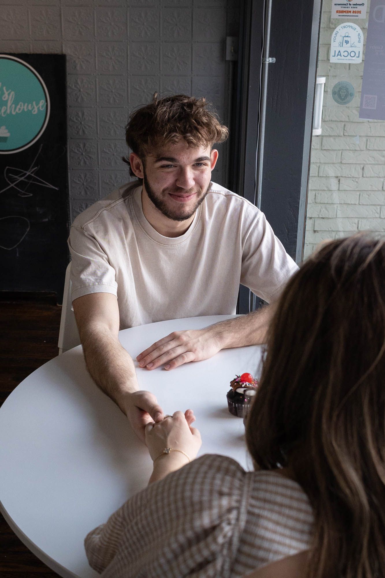 Smiling young man in white t-shirt holds hands with woman across a round white cafe table.