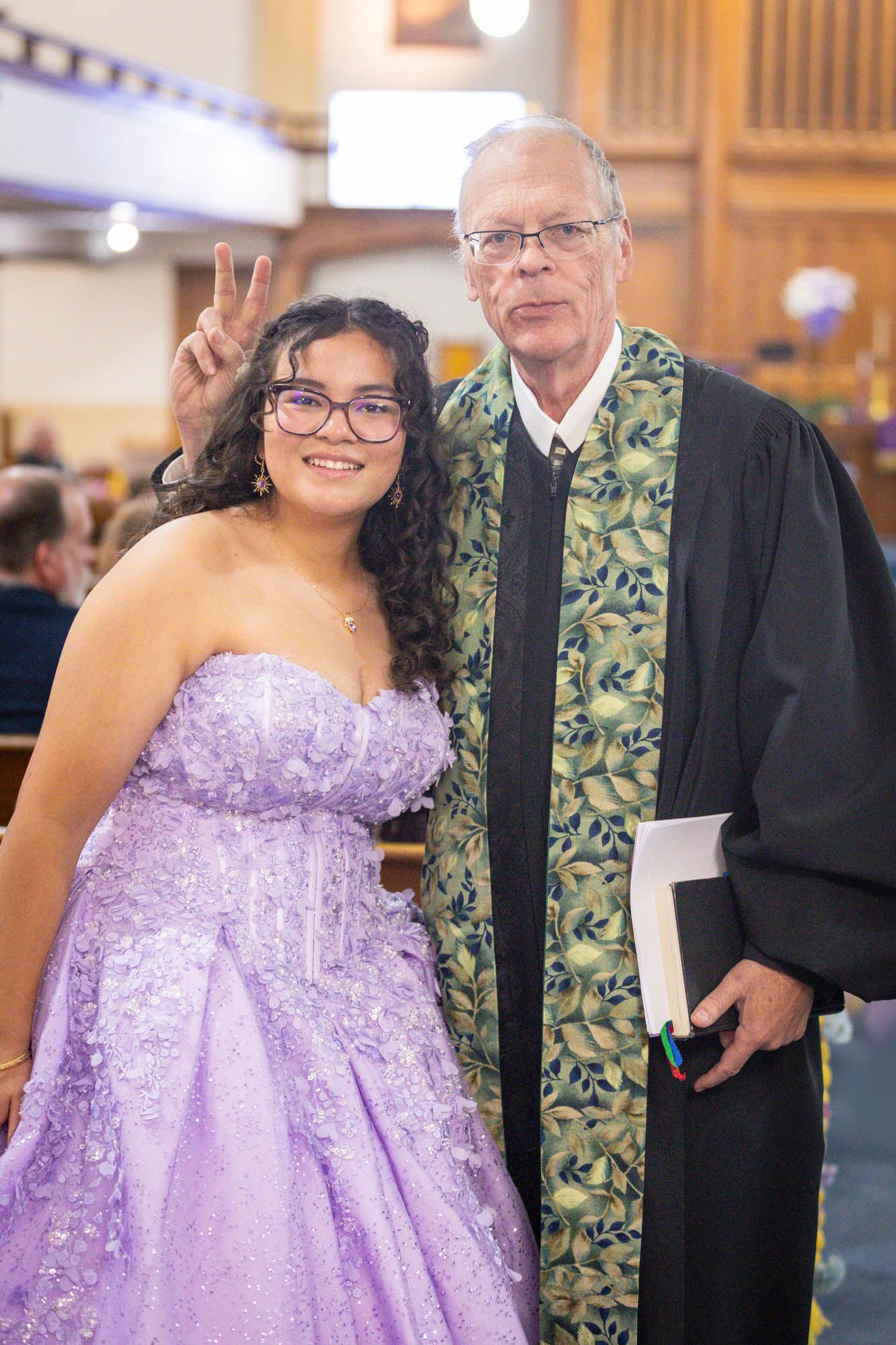 Young woman in lavender quinceañera gown poses with pastor in black robe inside a church.