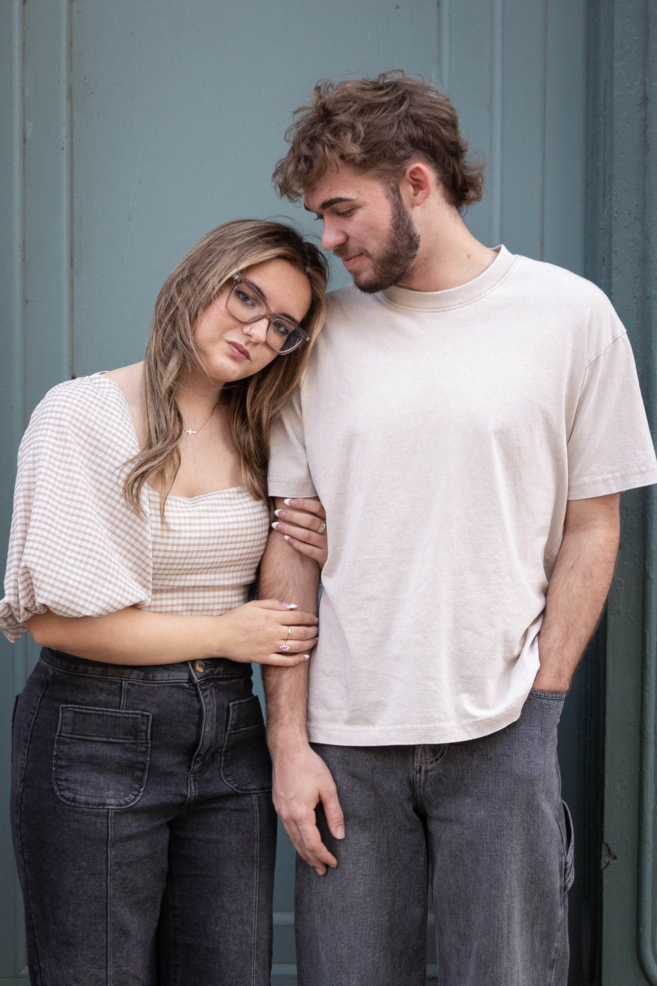 Young couple in casual outfits posing together, woman in glasses leaning on man's shoulder against teal wall.