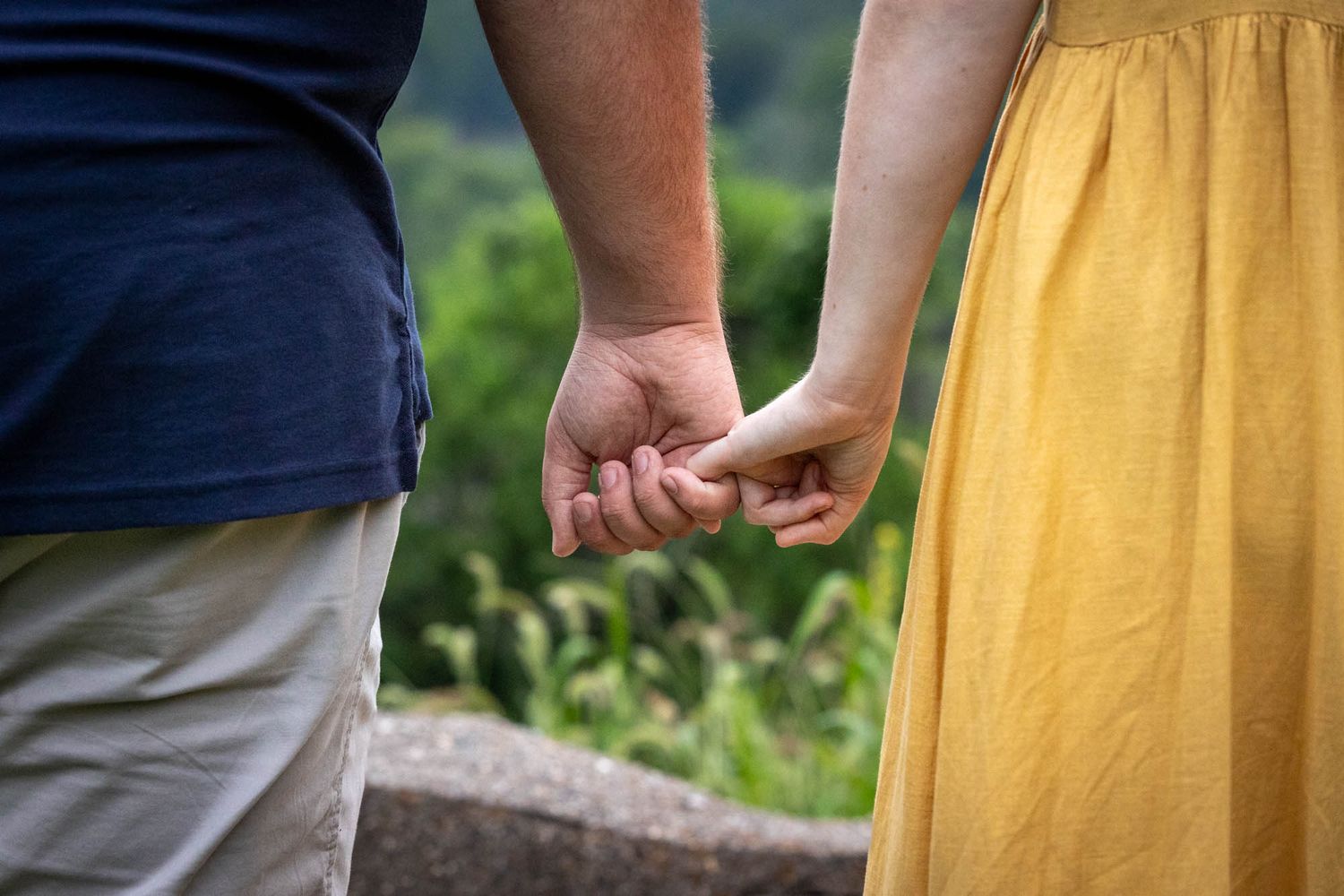 Couple holding hands outdoors, man in blue shirt and woman in yellow dress, with green nature background.