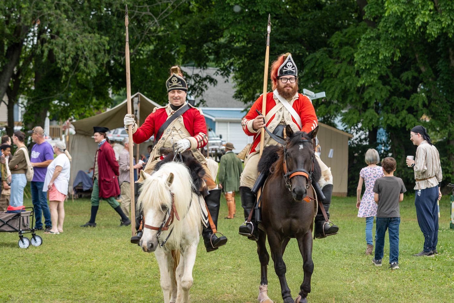 Two historical reenactors in red British colonial uniforms ride horses through a crowded outdoor festival grounds.