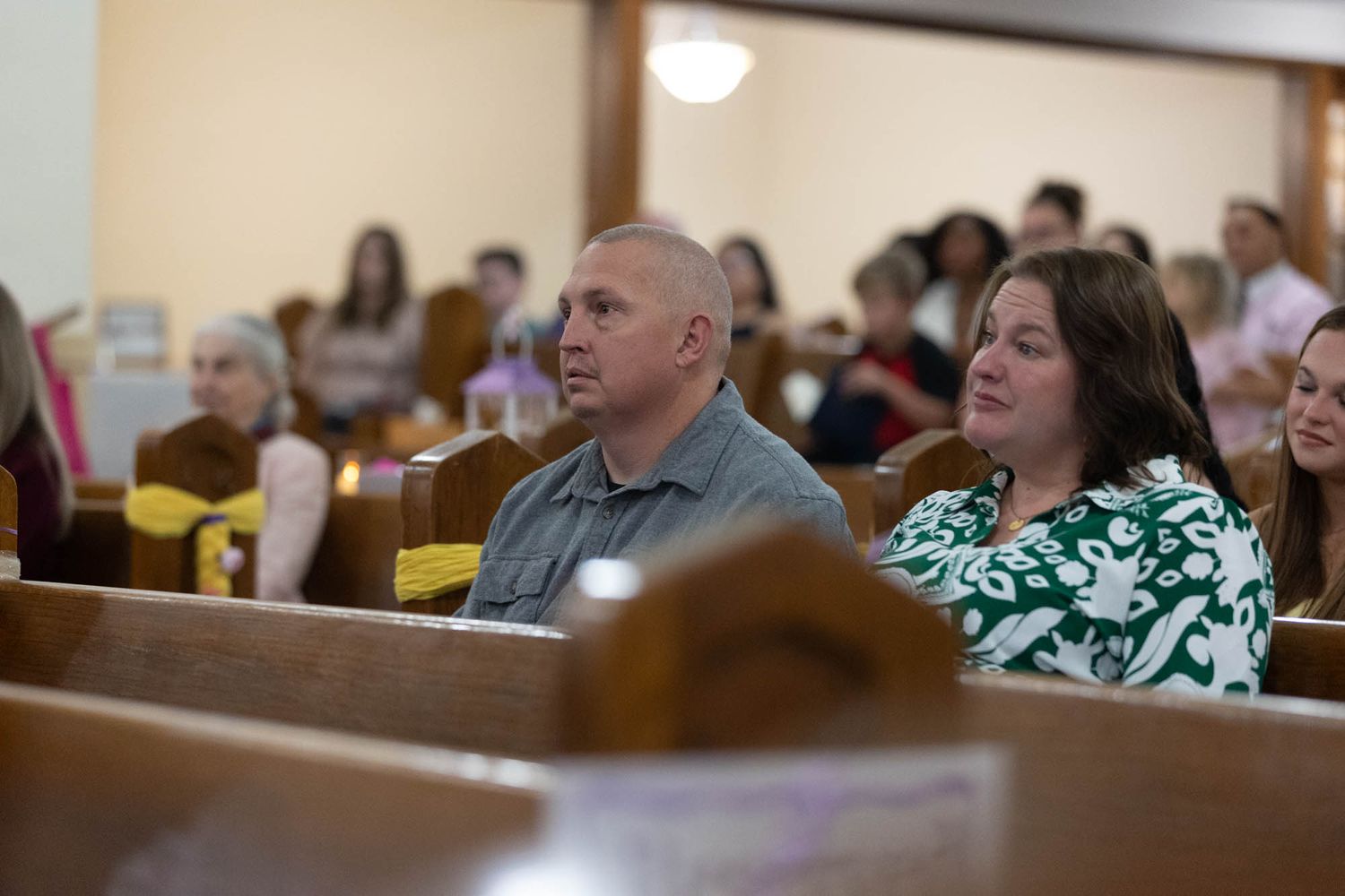 Guests seated in wooden church pews during a quinceañera ceremony, including a man and woman in the foreground.