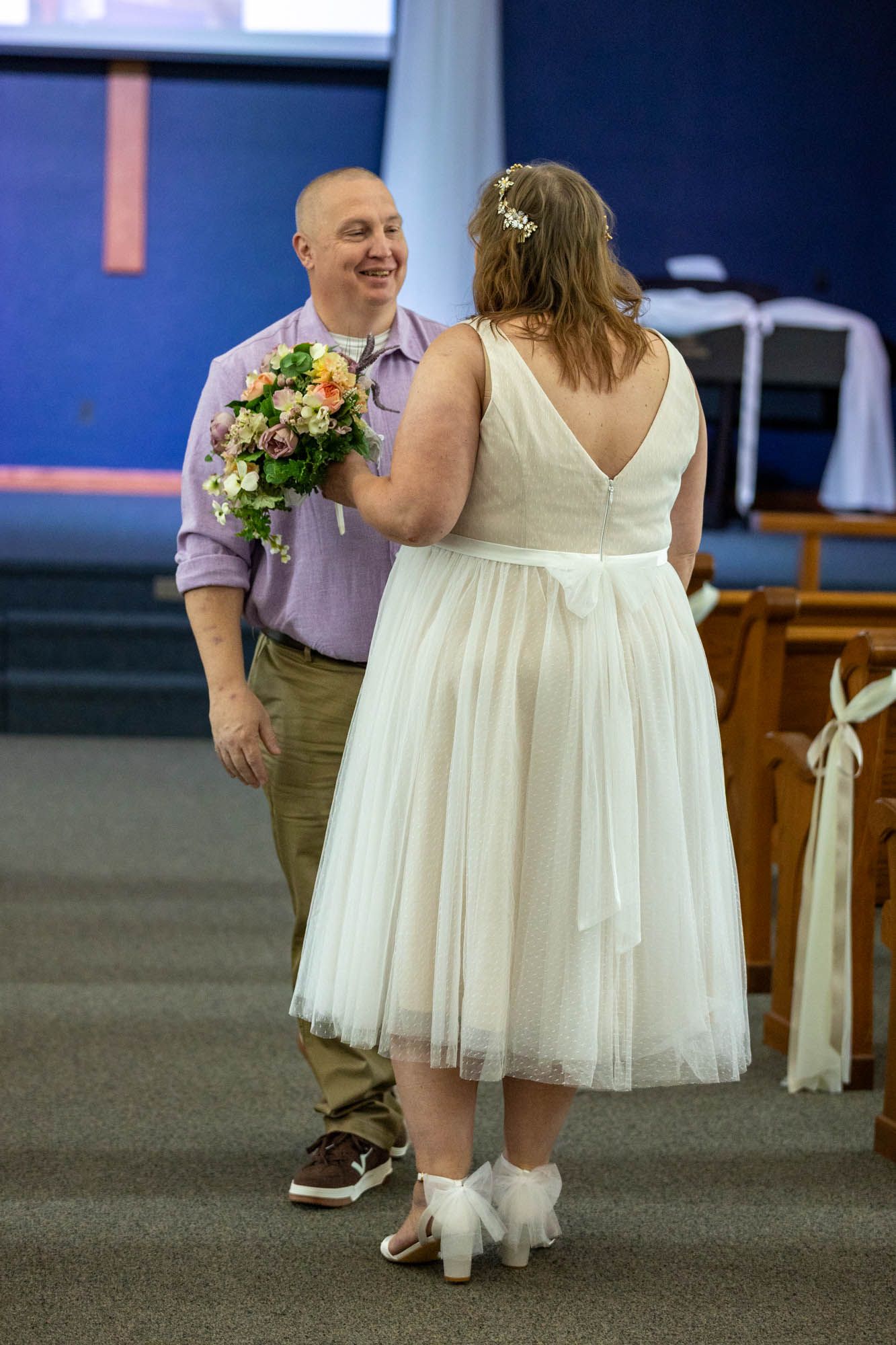 Bride in white tea-length dress holds bouquet facing smiling groom in lavender shirt during indoor wedding ceremony.