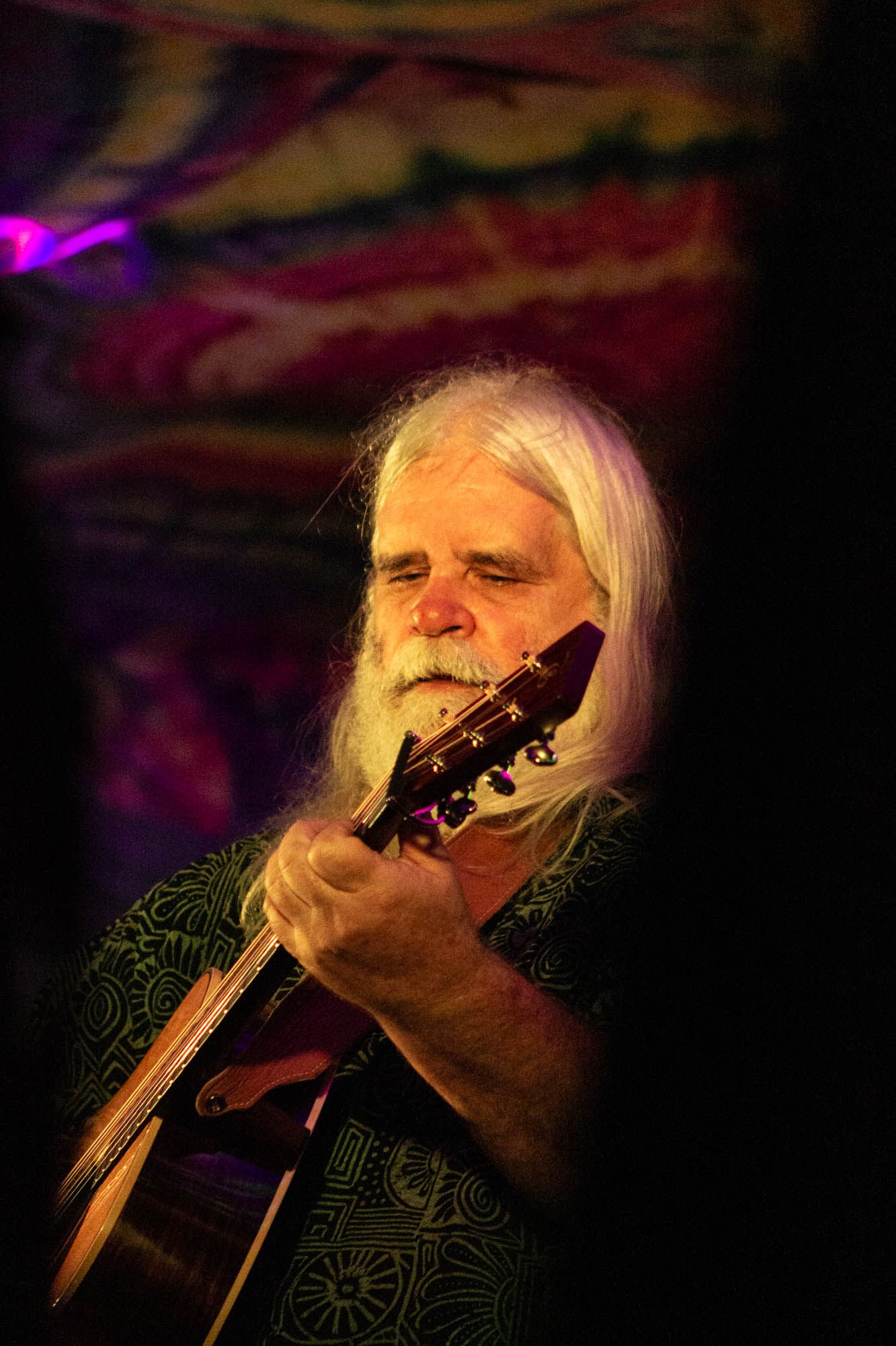 Long white-haired bearded man plays acoustic guitar on stage under purple and gold lights.
