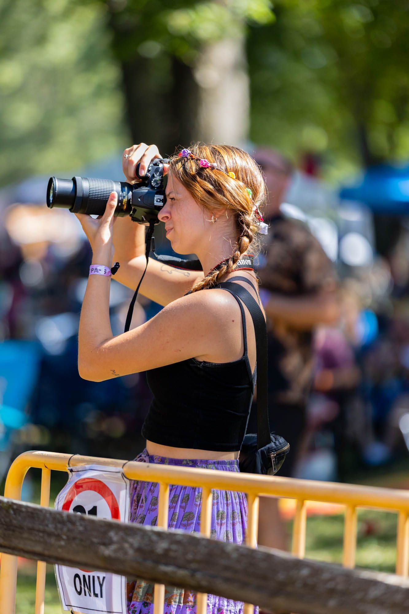 A young woman with a braided ponytail photographs an outdoor festival with a professional DSLR camera.