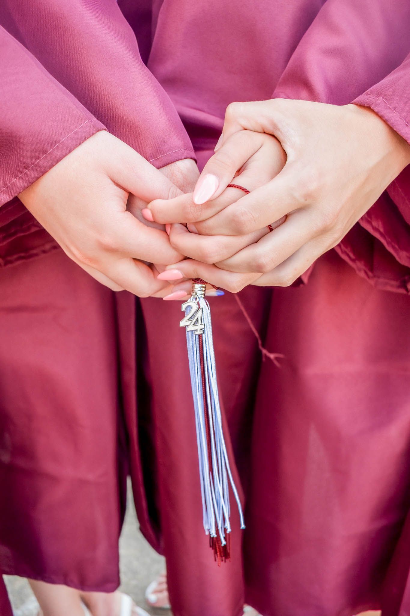 Close-up of graduates hands clasped together holding a silver tassel charm while wearing maroon graduation gowns.
