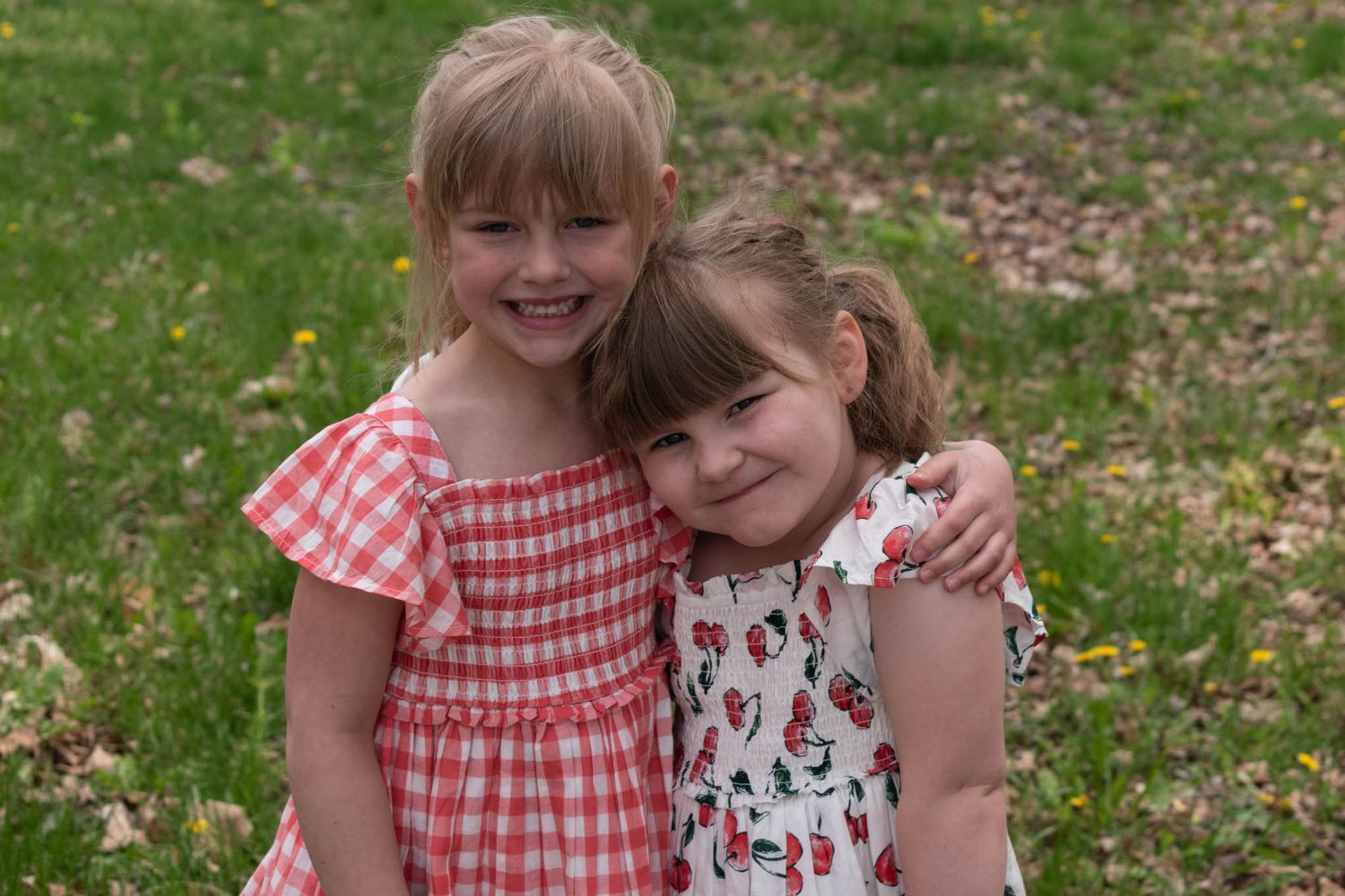 Two smiling little girls in red and white dresses embrace each other in a grassy outdoor setting.