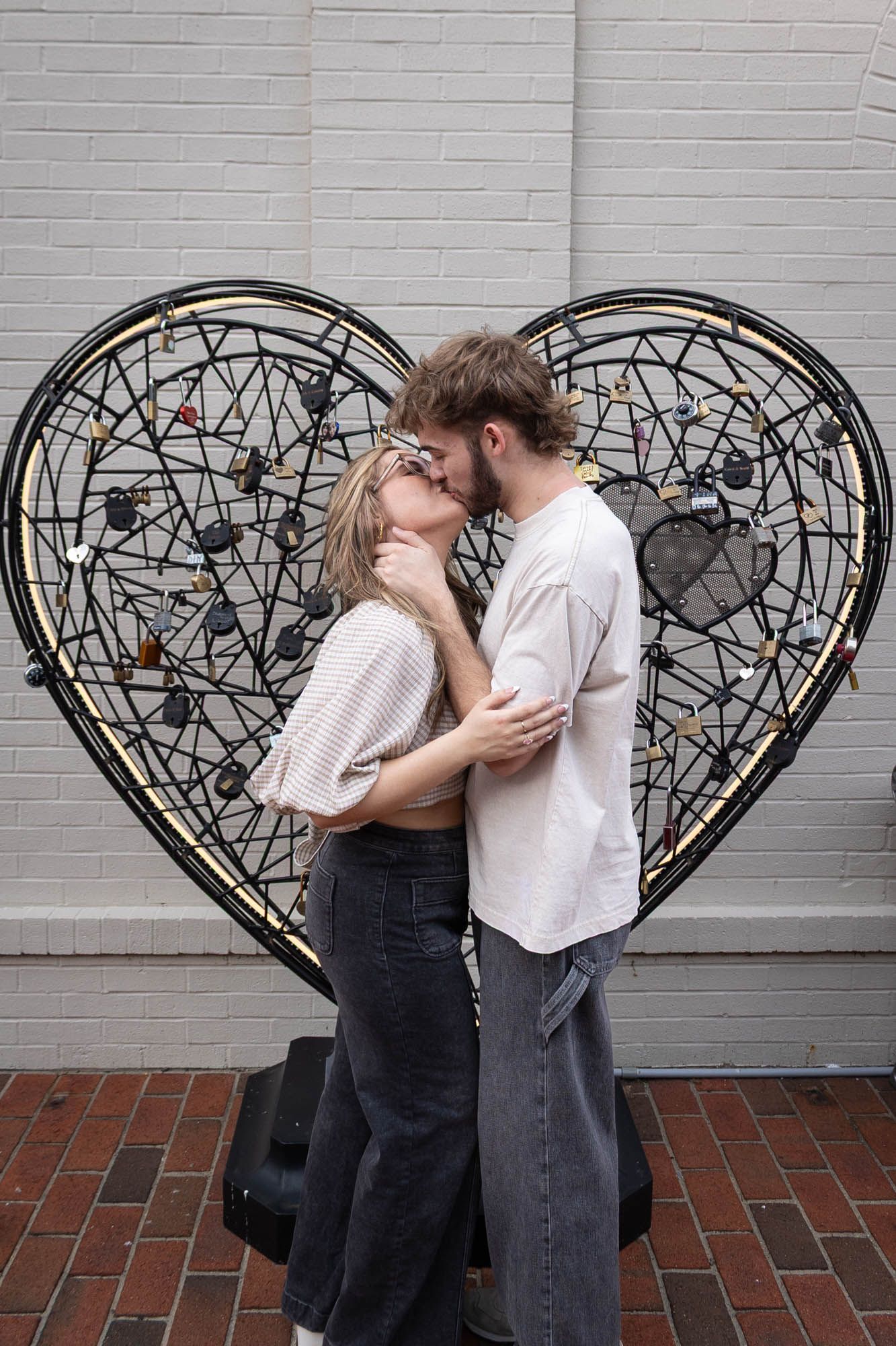 Couple kissing in front of a large black metal heart sculpture with love locks on a brick wall.