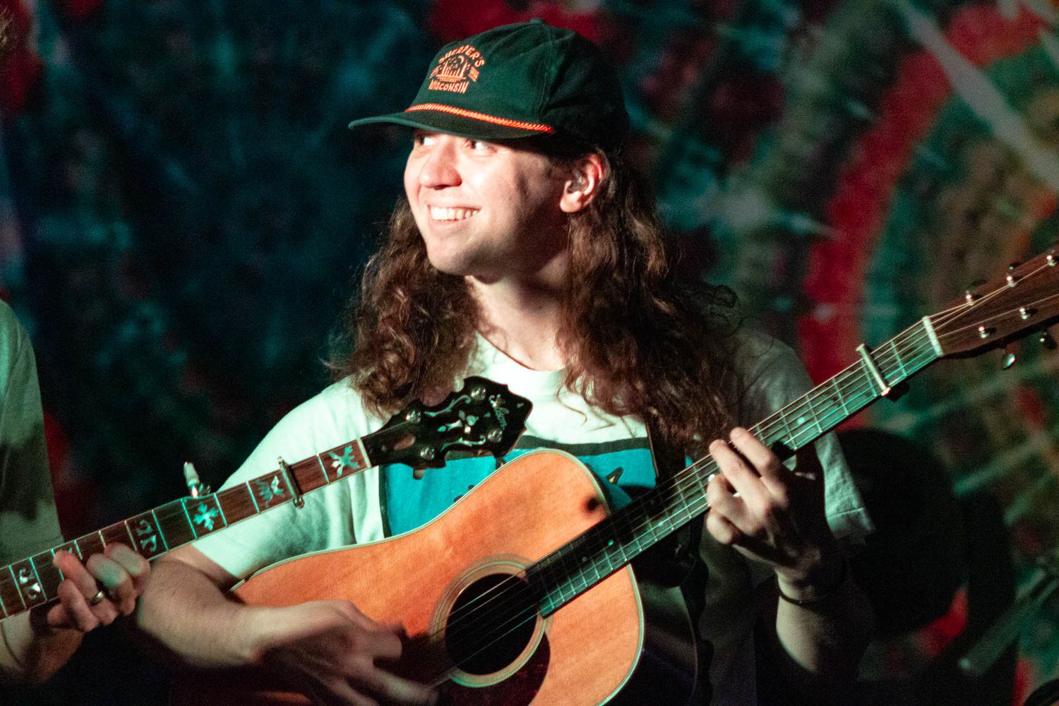 Smiling young musician with long hair plays acoustic guitar on stage with tie-dye backdrop.