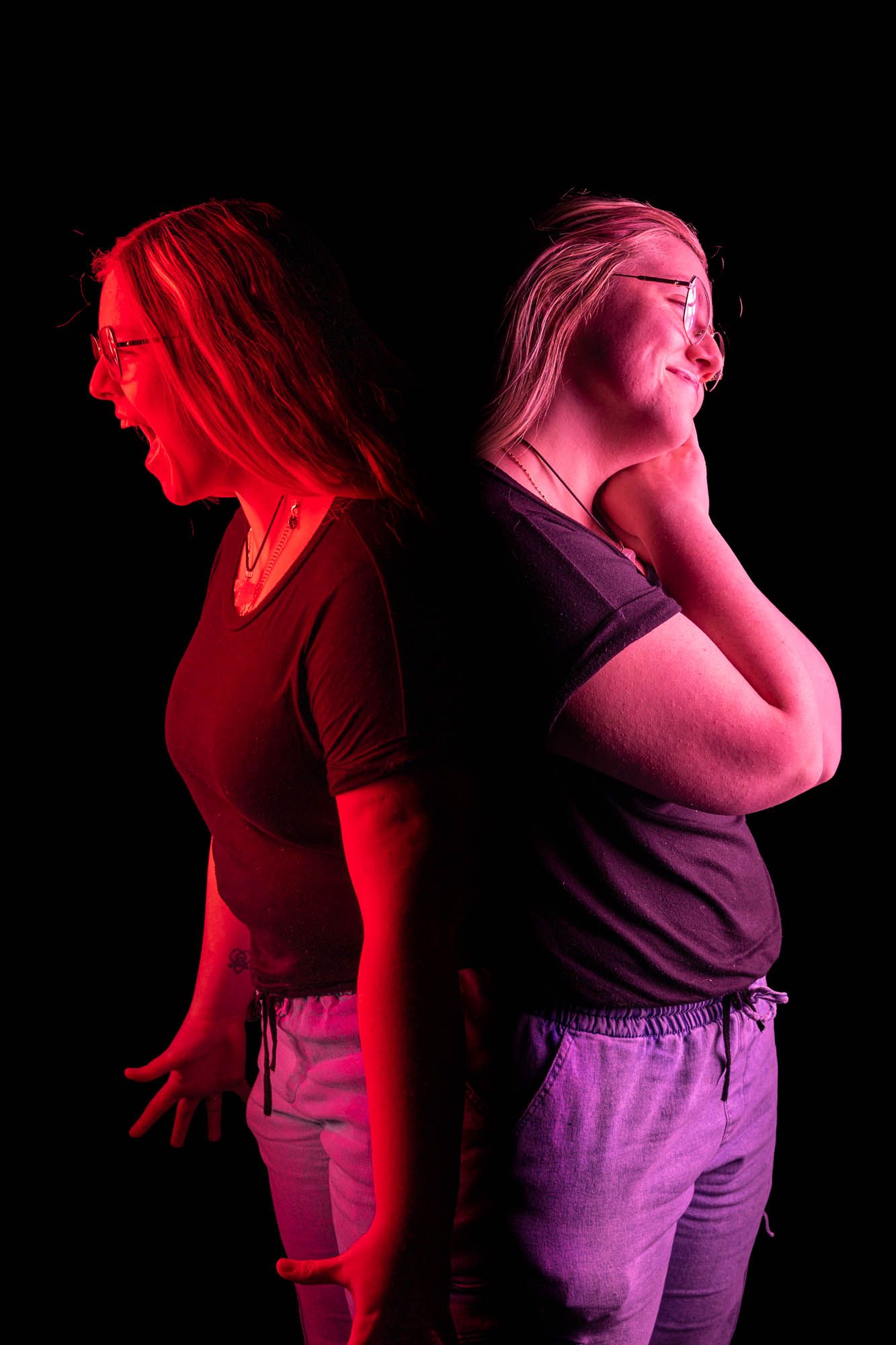 Two women back-to-back under dramatic red and purple lighting against black background, one laughing one screaming.