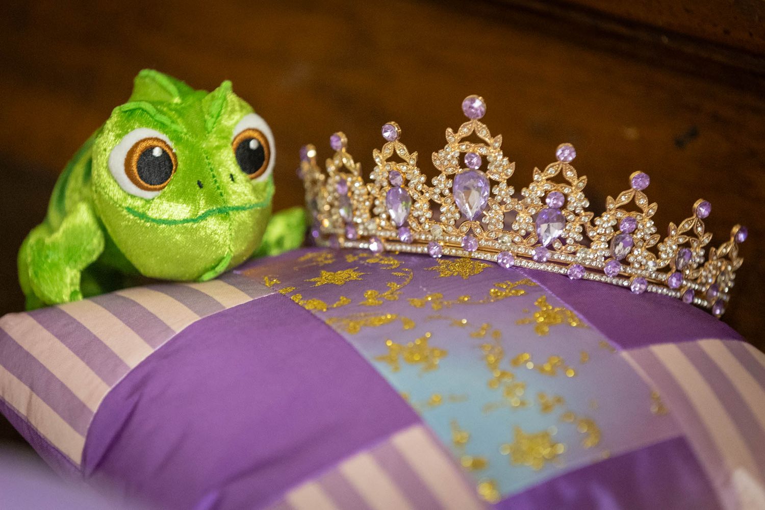 Pascal frog plush toy beside a purple gemstone tiara on a striped pillow.