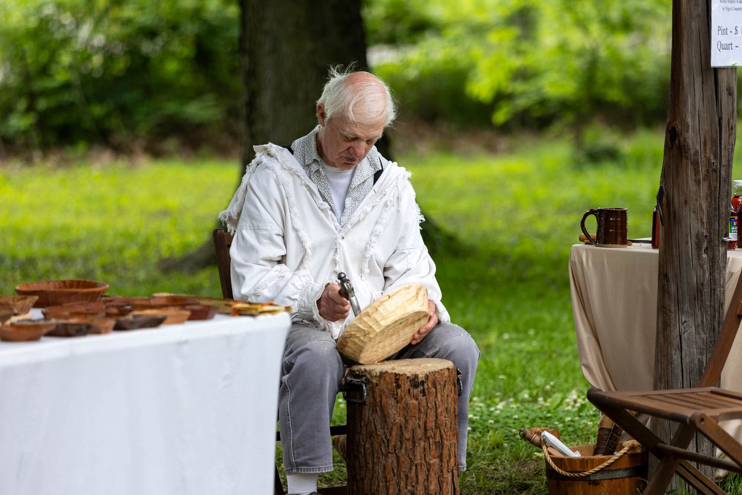 An elderly man carves wood while seated outdoors at a colonial crafts demonstration at a historical festival.