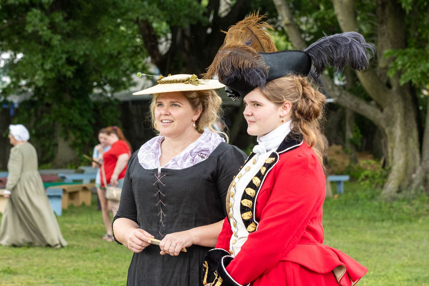 Two women in elegant 18th century period costumes with feathered hats at outdoor reenactment.